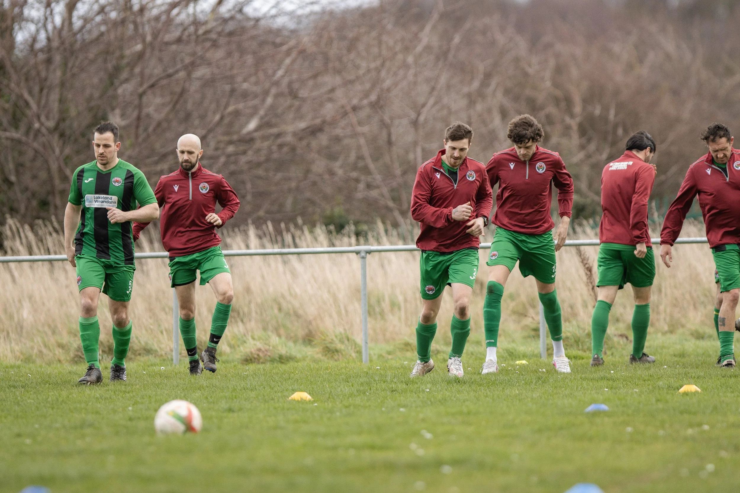 Soccer players warming up on the field during a training session, wearing green shorts and red and green warm-up jackets.