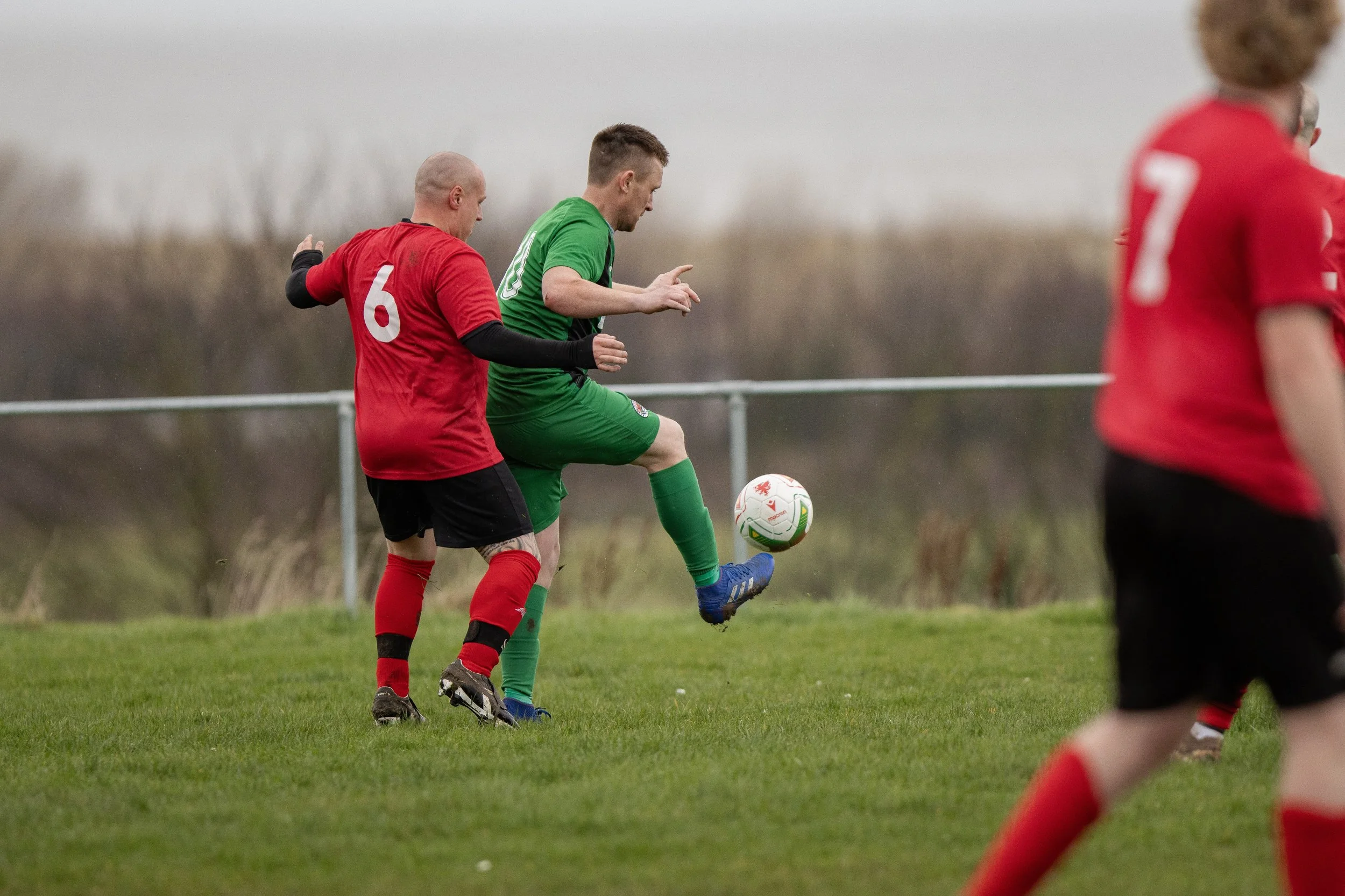 Soccer players competing for the ball on the field during a game, one in a red uniform and another in a green uniform, with a grassy field and a cloudy sky in the background.