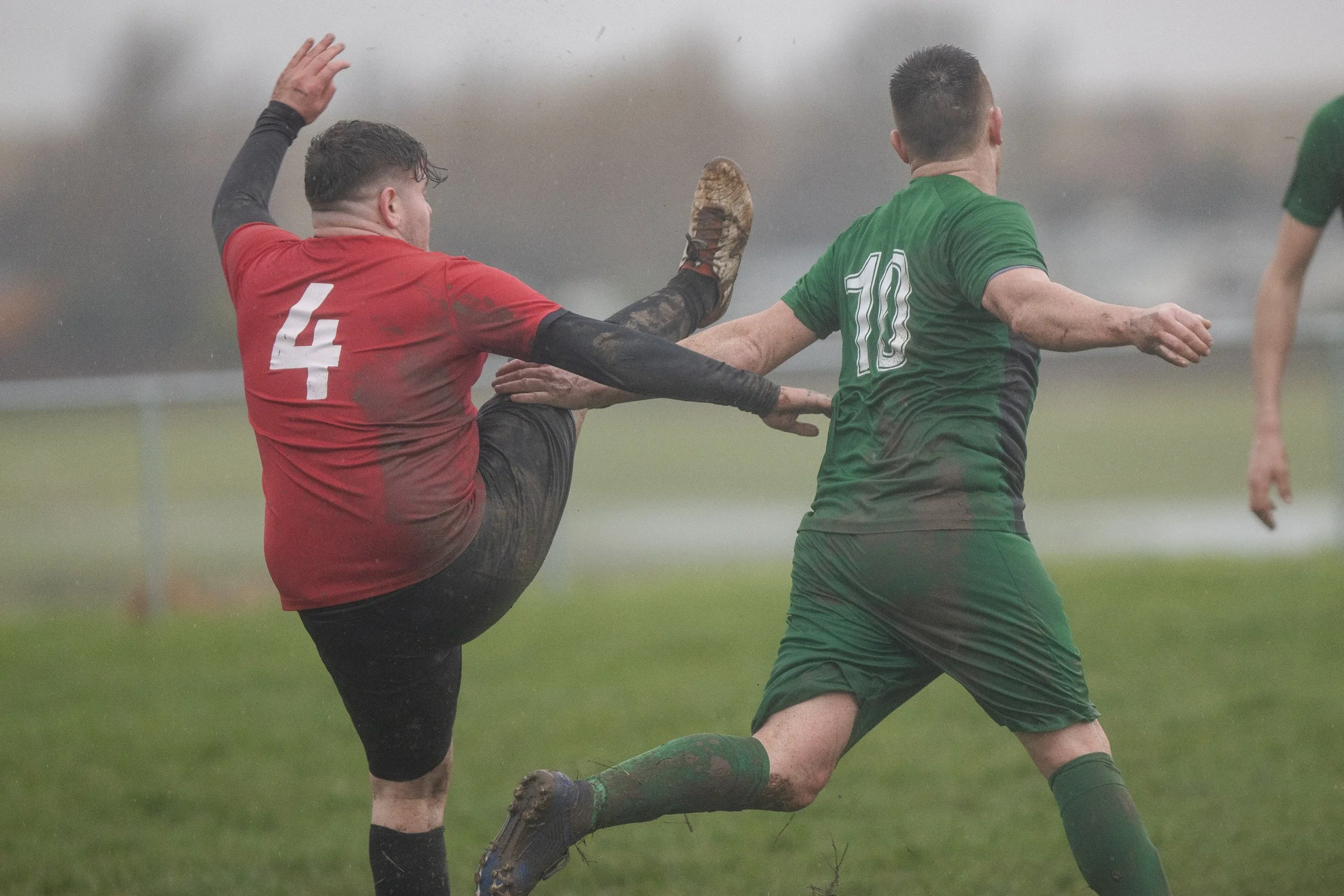 Two soccer players in muddy uniforms competing for the ball on a rainy day. One is wearing a red jersey with the number 4, and the other is wearing a green jersey with the number 10.