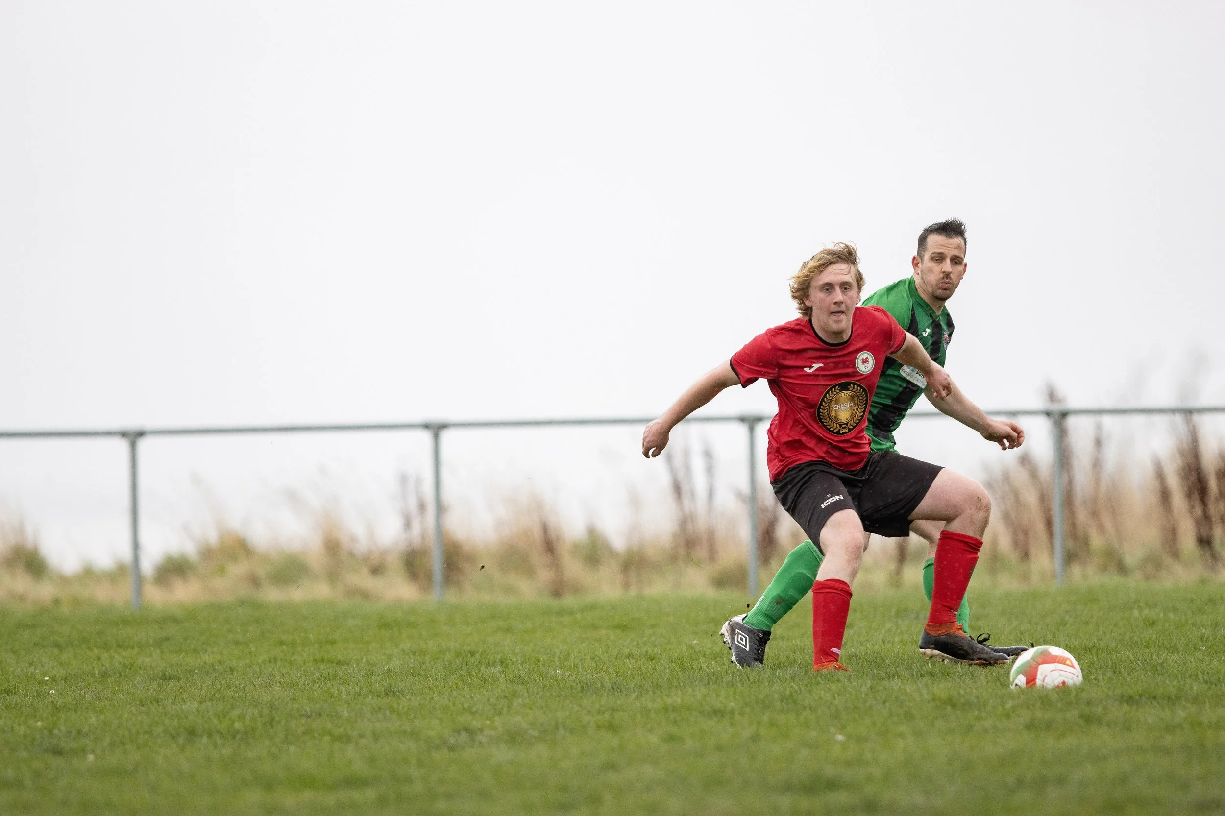 Two soccer players, one in a red jersey and the other in a green jersey, compete for the ball on a grassy field during daytime. The player in red is slightly in front, with the player in green closely behind, both focused on the ball.