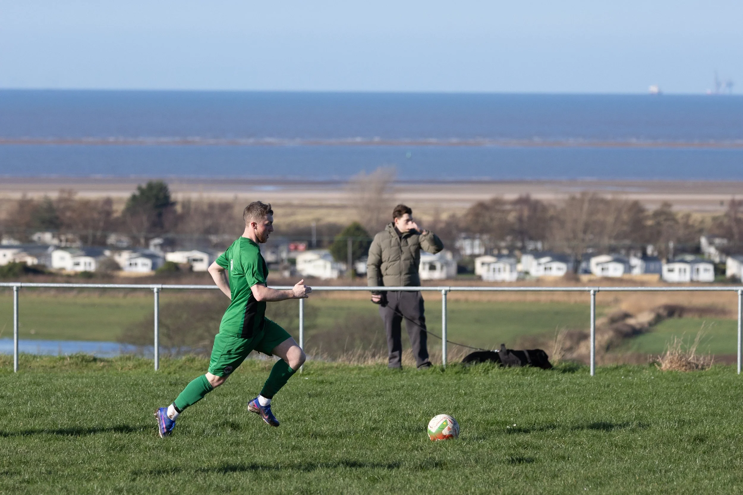 A man in a green soccer uniform running on a grassy field with a soccer ball, while a person in a brown jacket watches from behind a fence, with a background of houses, trees, a body of water, and a blue sky.