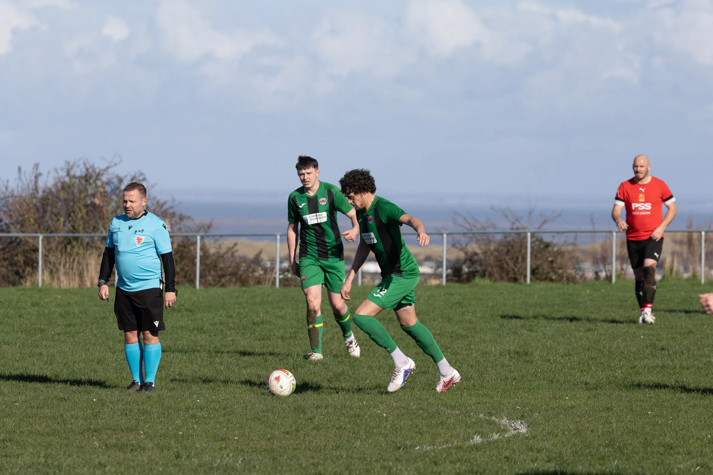 A soccer match on a grassy field with four players and a referee. One player is about to kick the ball, while the others observe. The players are wearing different colored jerseys, and there is a fence and trees in the background under a partly cloud