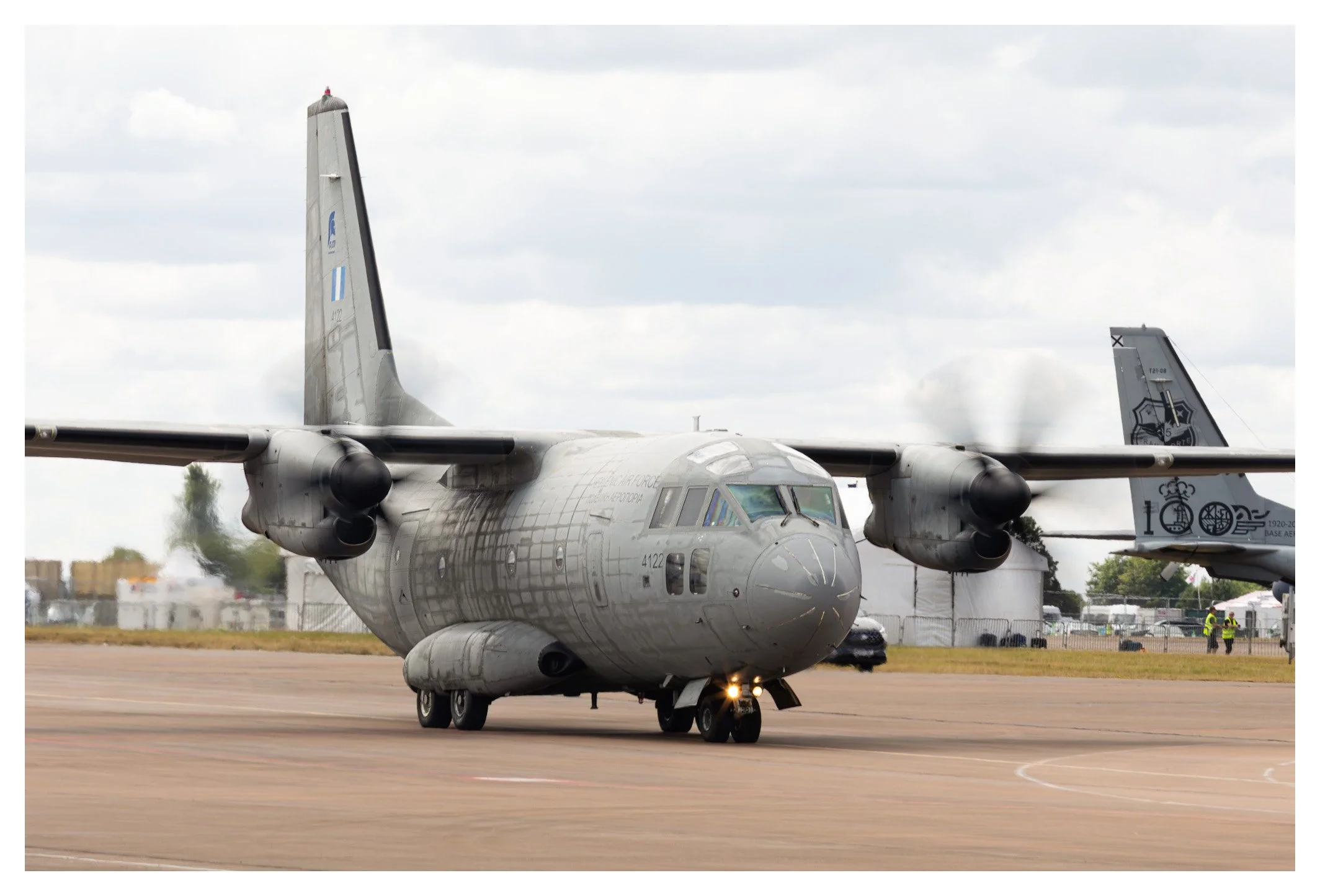 A military cargo airplane on the runway at an airfield during daytime, with other aircraft and personnel in the background.