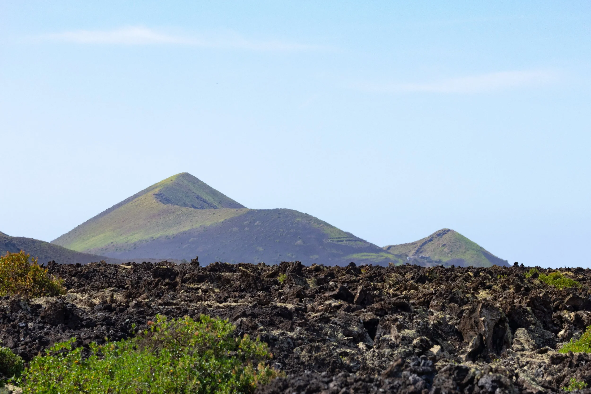 Volcanic landscape with dark lava rocks in the foreground and green-capped volcanic peaks in the background under a blue sky.