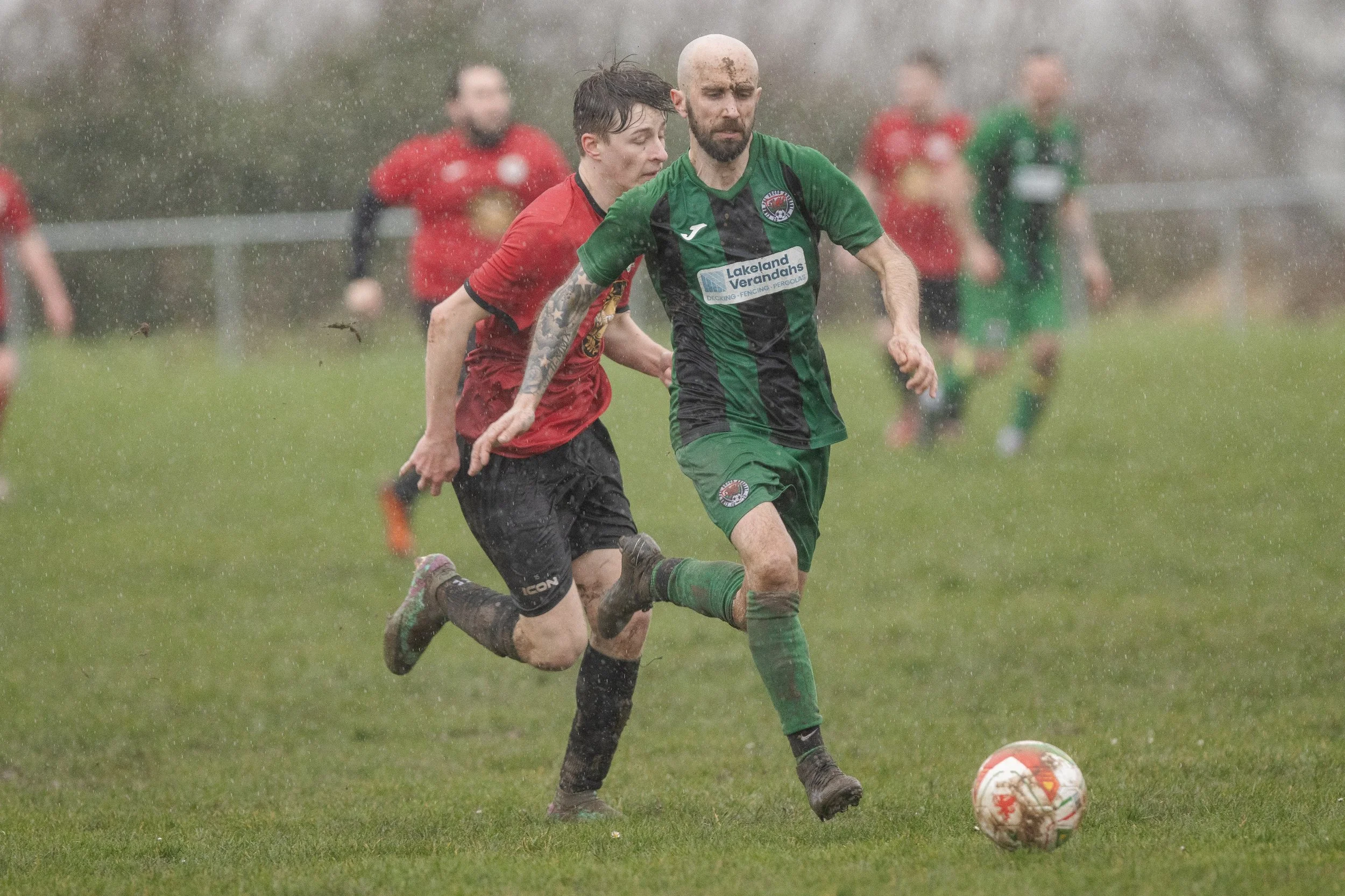 Two soccer players, one in red and black and the other in green and black, playing in the rain on a grassy field. The player in green is kicking the ball while the player in red is behind him. There are more players blurred in the background.