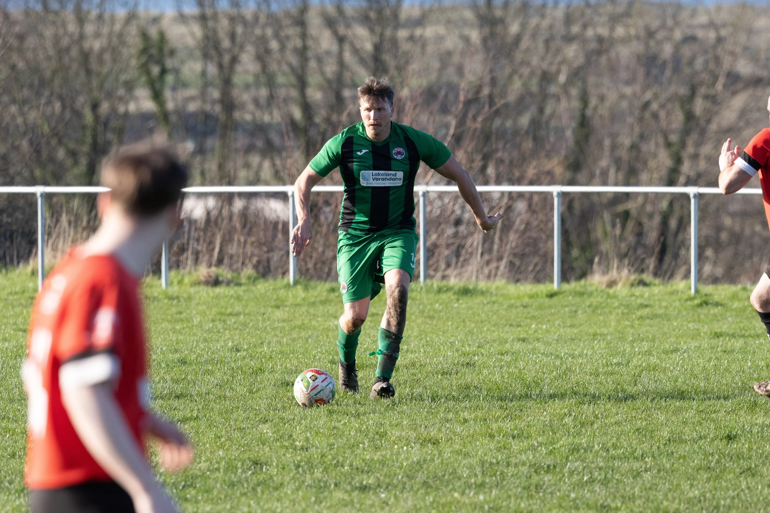 A soccer player in a green and black uniform is dribbling the ball on a grass field during a match, with other players in red and black uniforms visible in the foreground and background.