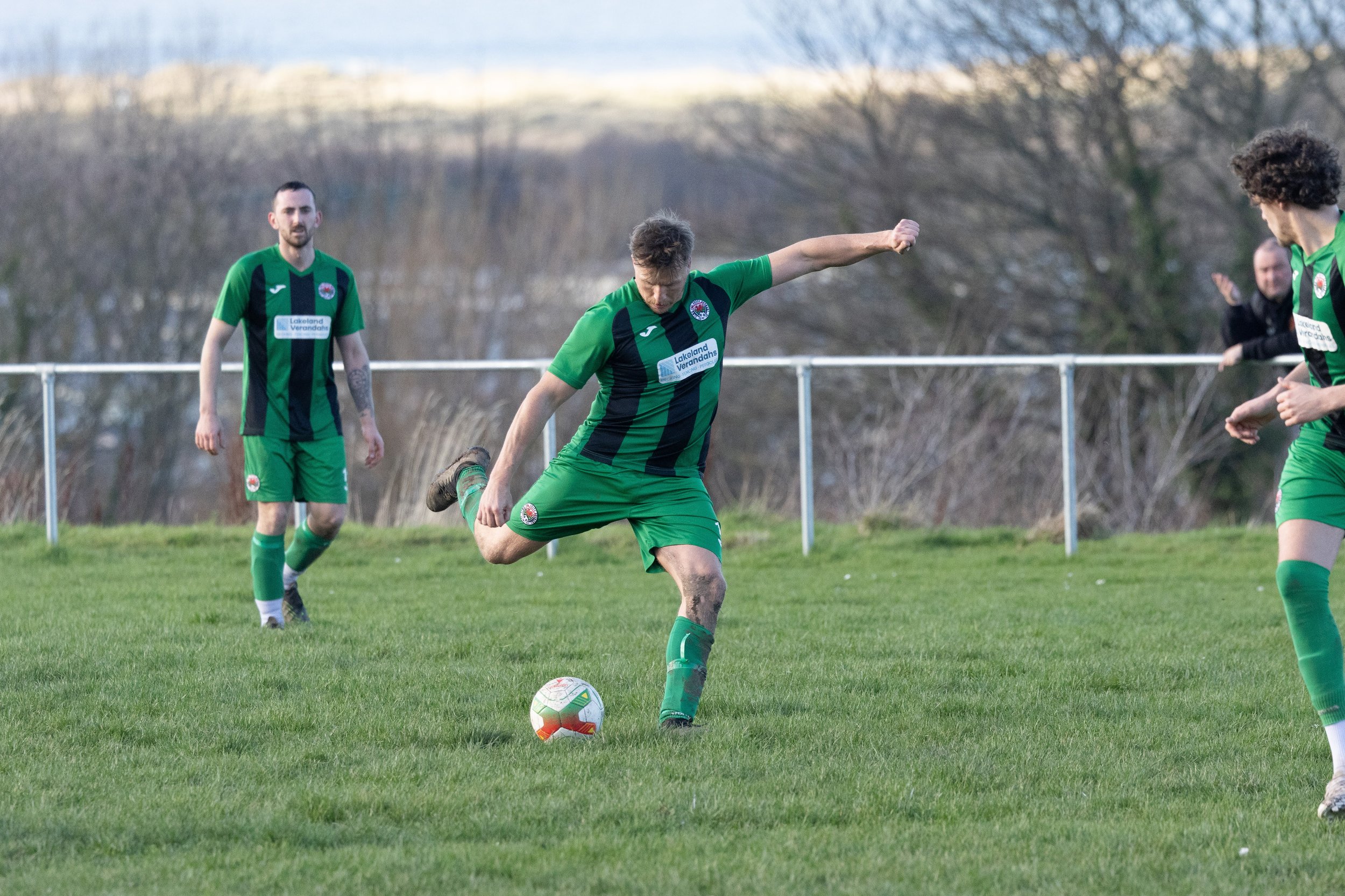 A soccer player in green and black uniform kicking a ball on a grassy field, with two other players and a spectator in the background.