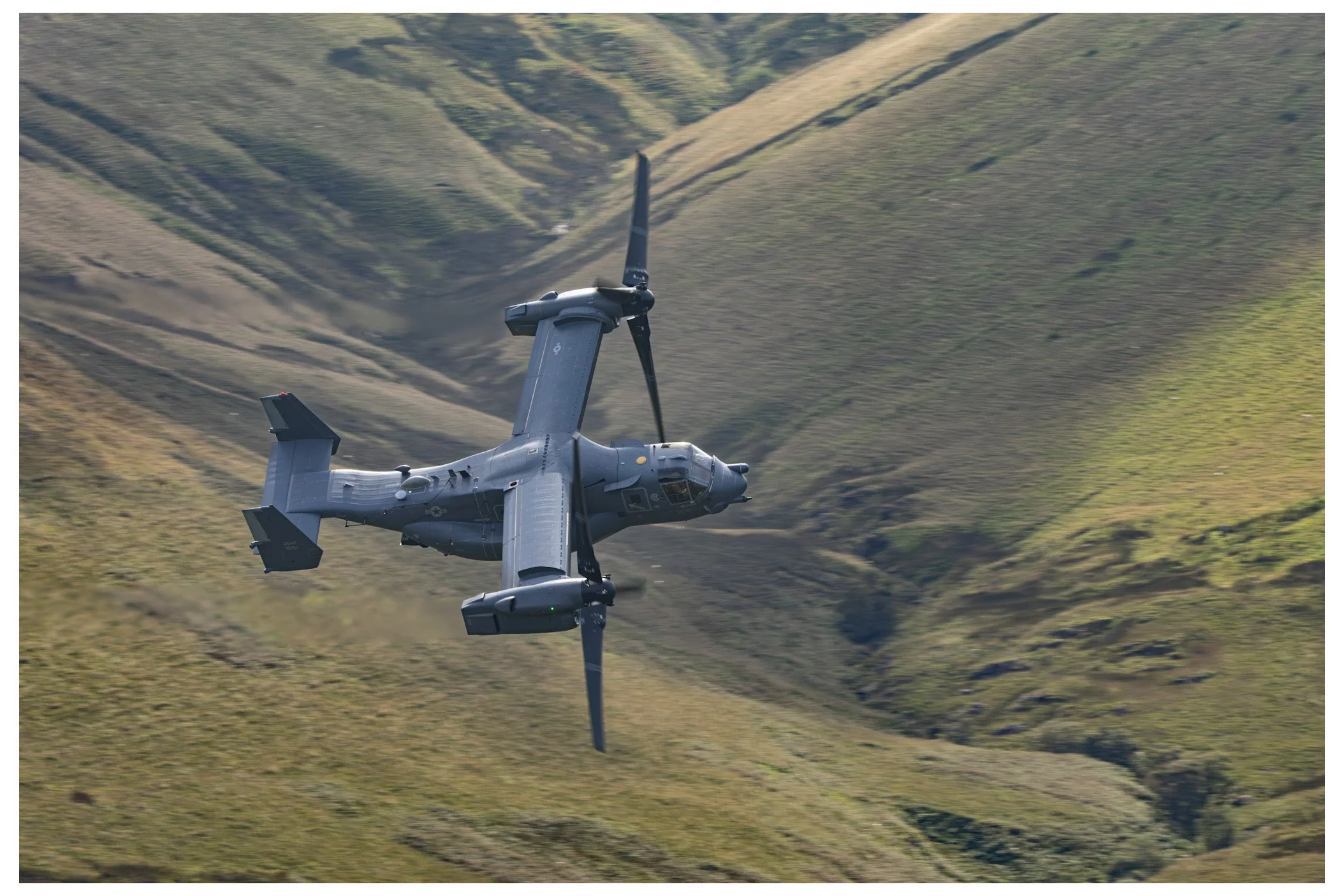 A gray military tiltrotor aircraft flying over a green hilly landscape with multiple ridges.