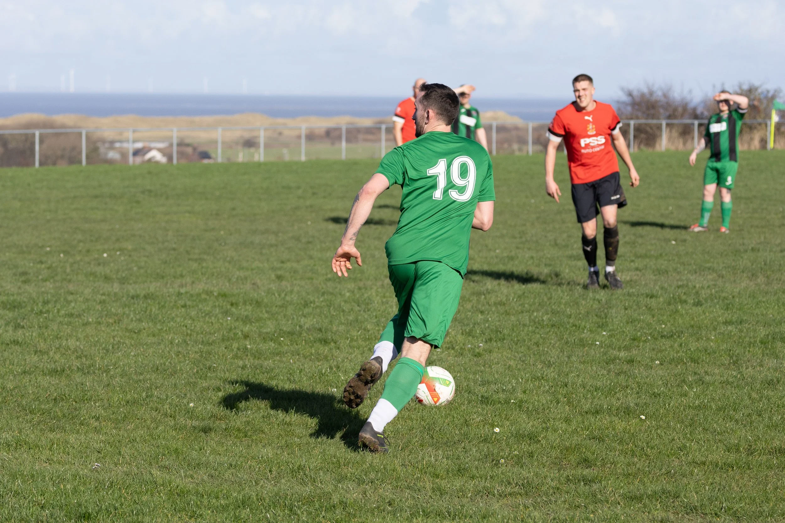 A soccer player in a green uniform with the number 19, kicking a soccer ball on a grassy field. Four other players are in the background, some in green, some in red, with a fence and a landscape with wind turbines and water in the distance.