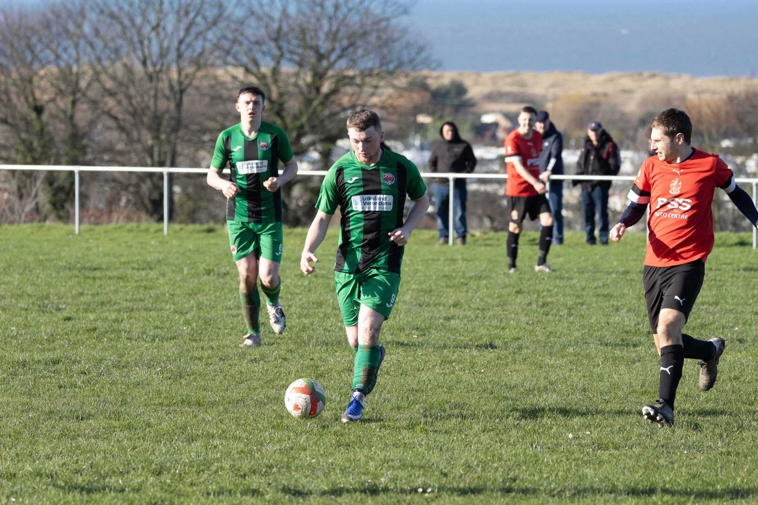 Soccer players on the field, with one in green about to kick the ball, and others in the background watching, on a grassy field with a scenic backdrop of trees and water.