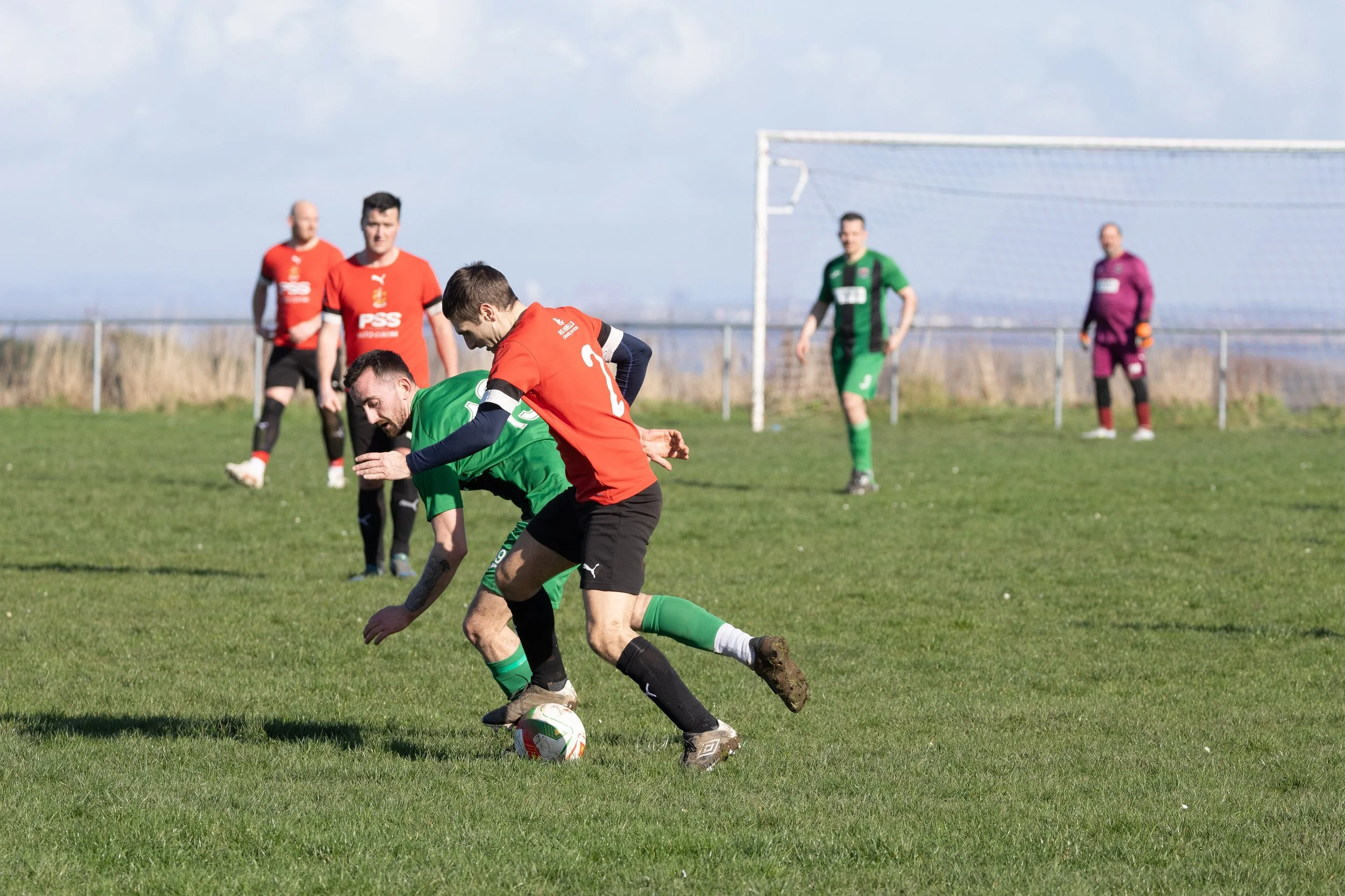 Two soccer players, one in a green uniform and the other in a red uniform, compete for the ball on a grassy field. Four other players and a goalie are visible in the background, with a goalpost and clear sky.