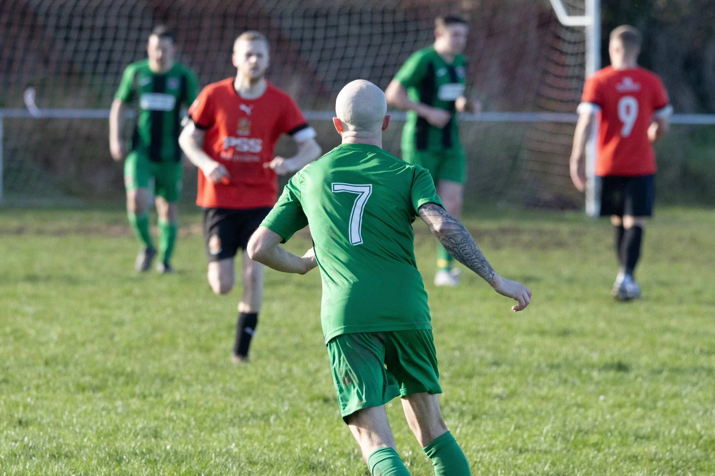 Soccer players on field, central player in green jersey with number 7, bald head, tattooed arm, close up.