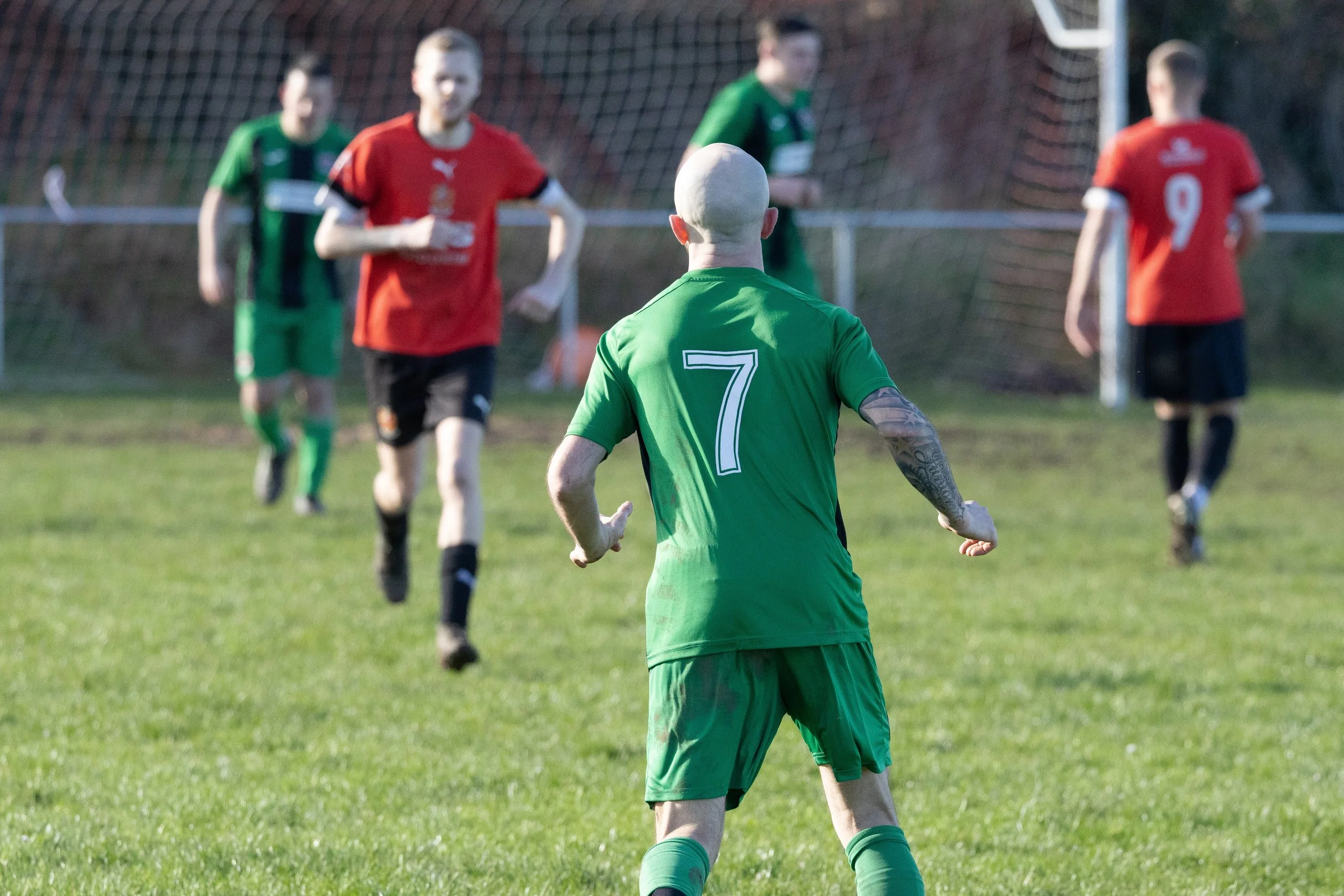 A soccer player with the number 7 on his green jersey is running towards the goal while players in red and green jerseys run in the background on a grassy soccer field.
