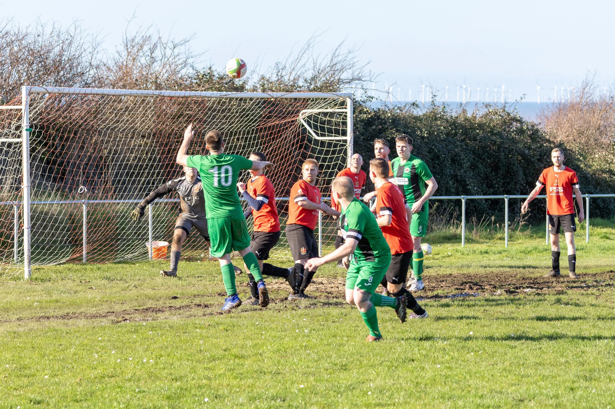 Soccer game with players in green and orange jerseys near goal, ball in air, and goalkeeper preparing to react.