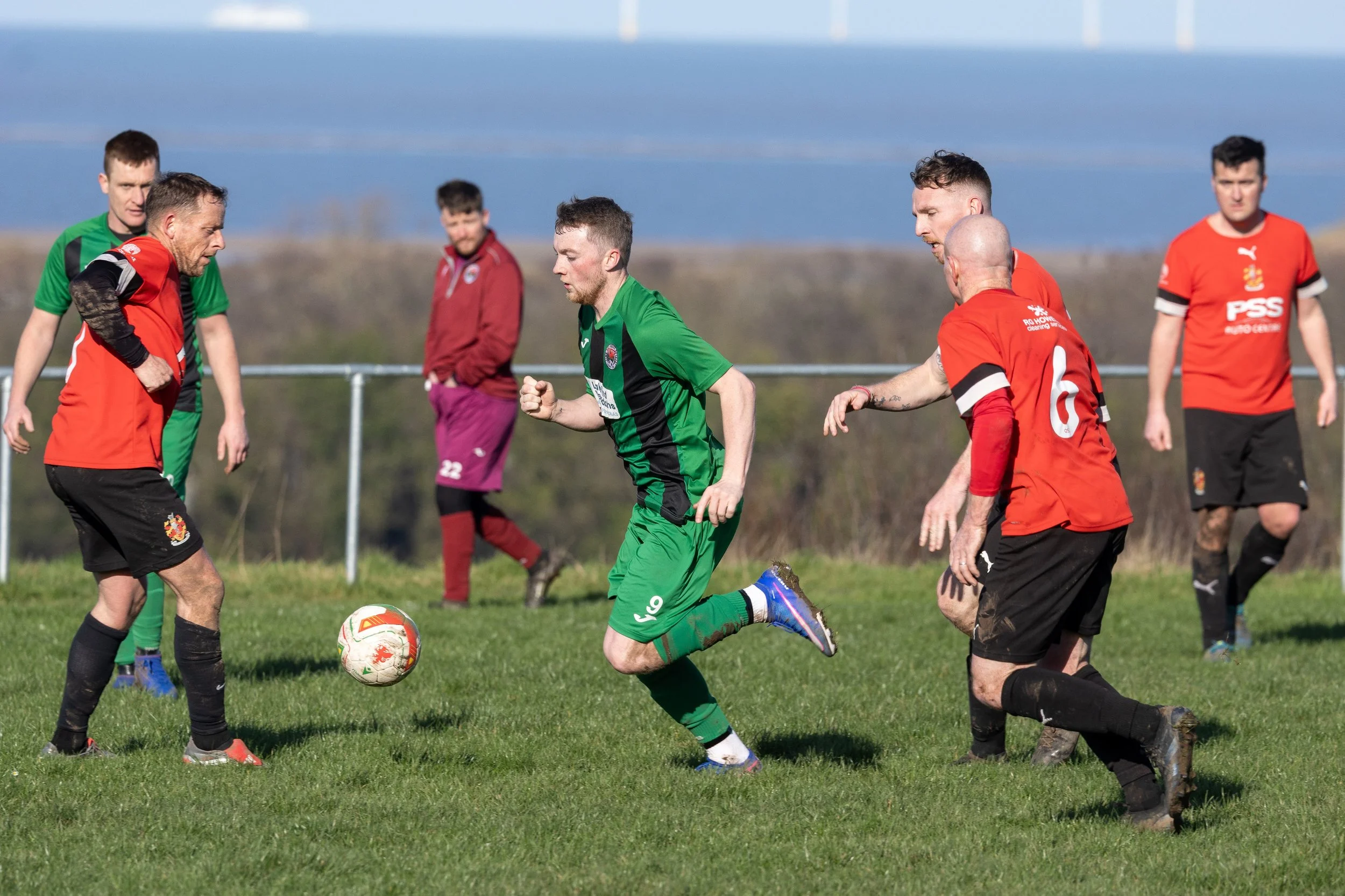 Soccer match with players in green and red jerseys on a grassy field, with a backdrop of trees and a blue sky.