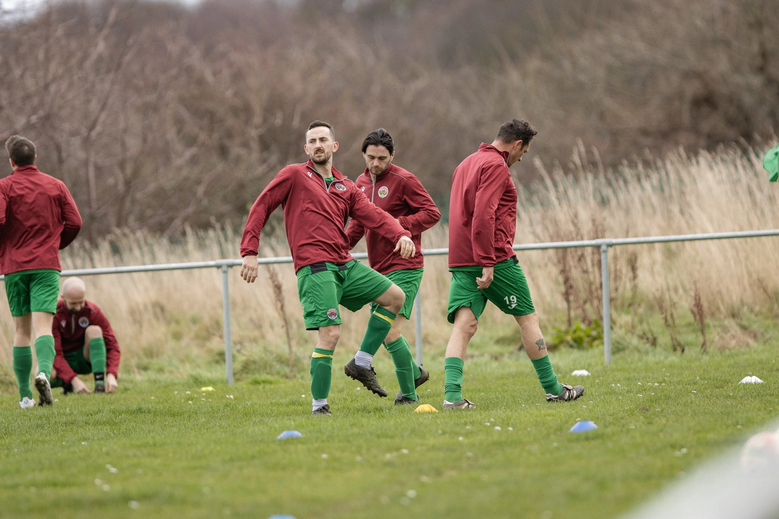 Soccer players in red jackets and green shorts warming up on a grassy field with cones, practicing stretching and warm-up exercises.