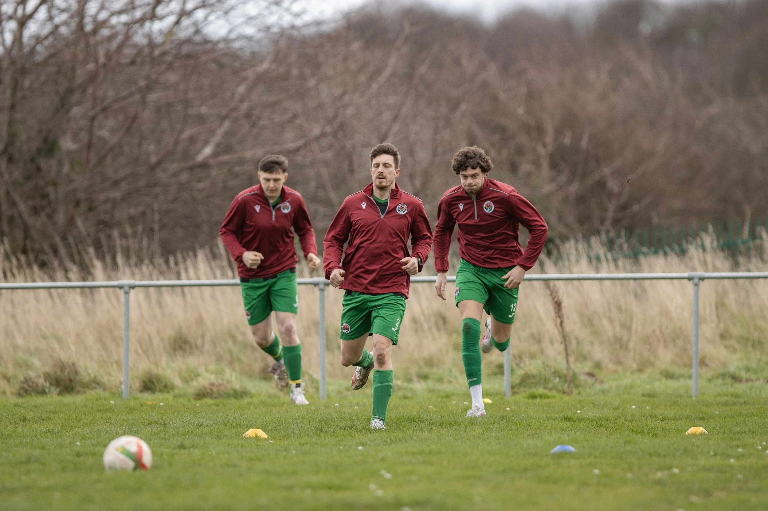 Three soccer players in maroon jackets and green shorts jogging on a grassy field during training.