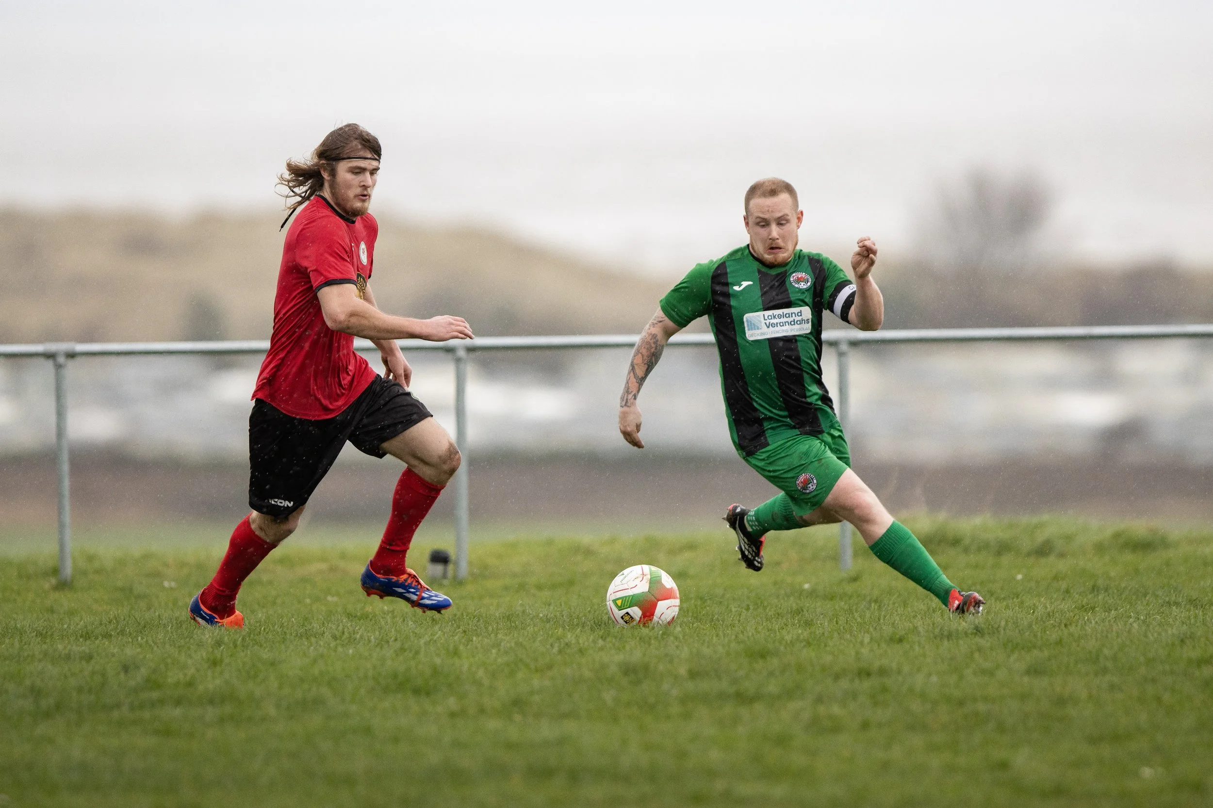 Two soccer players in action on a grassy field, one in a red uniform and the other in green, chasing the ball.