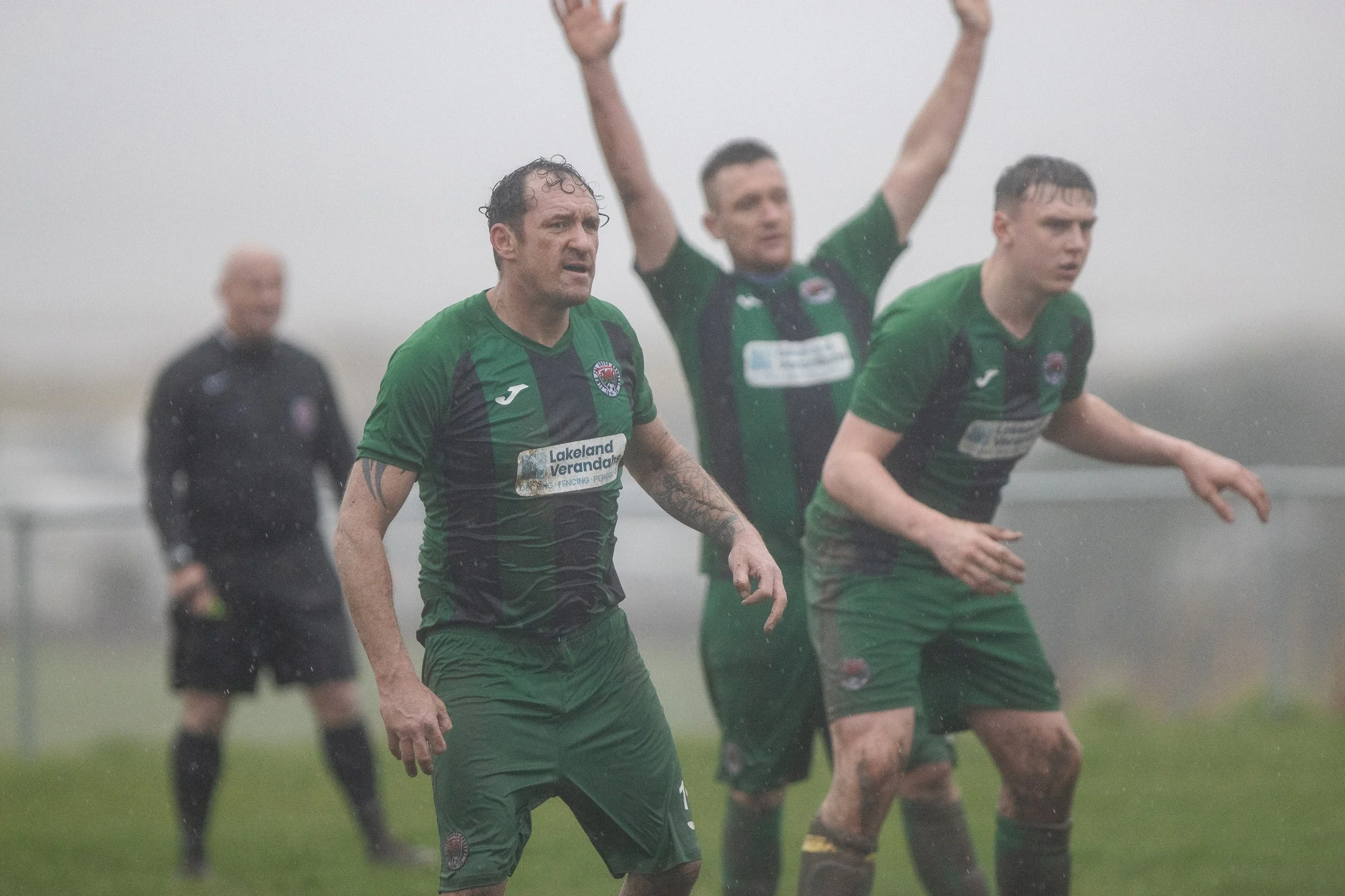 Four male soccer players in green and black uniforms on a rainy field, with one official in the background, during a match.