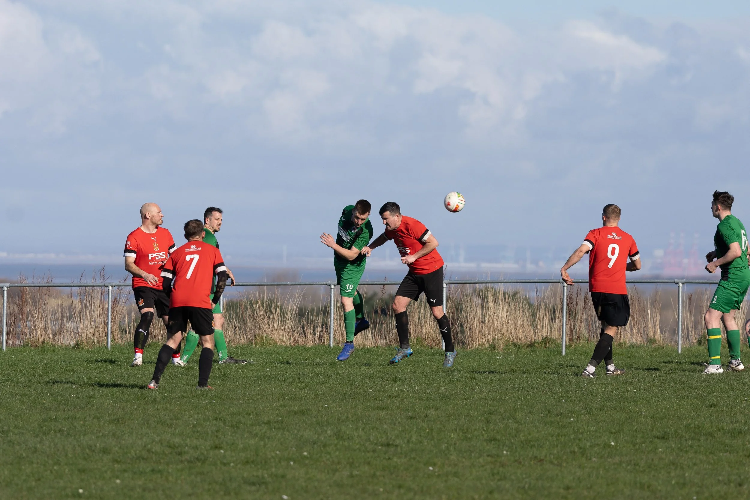 Soccer players in red and green jerseys playing on a grassy field during daytime, with a blue sky and clouds in the background.