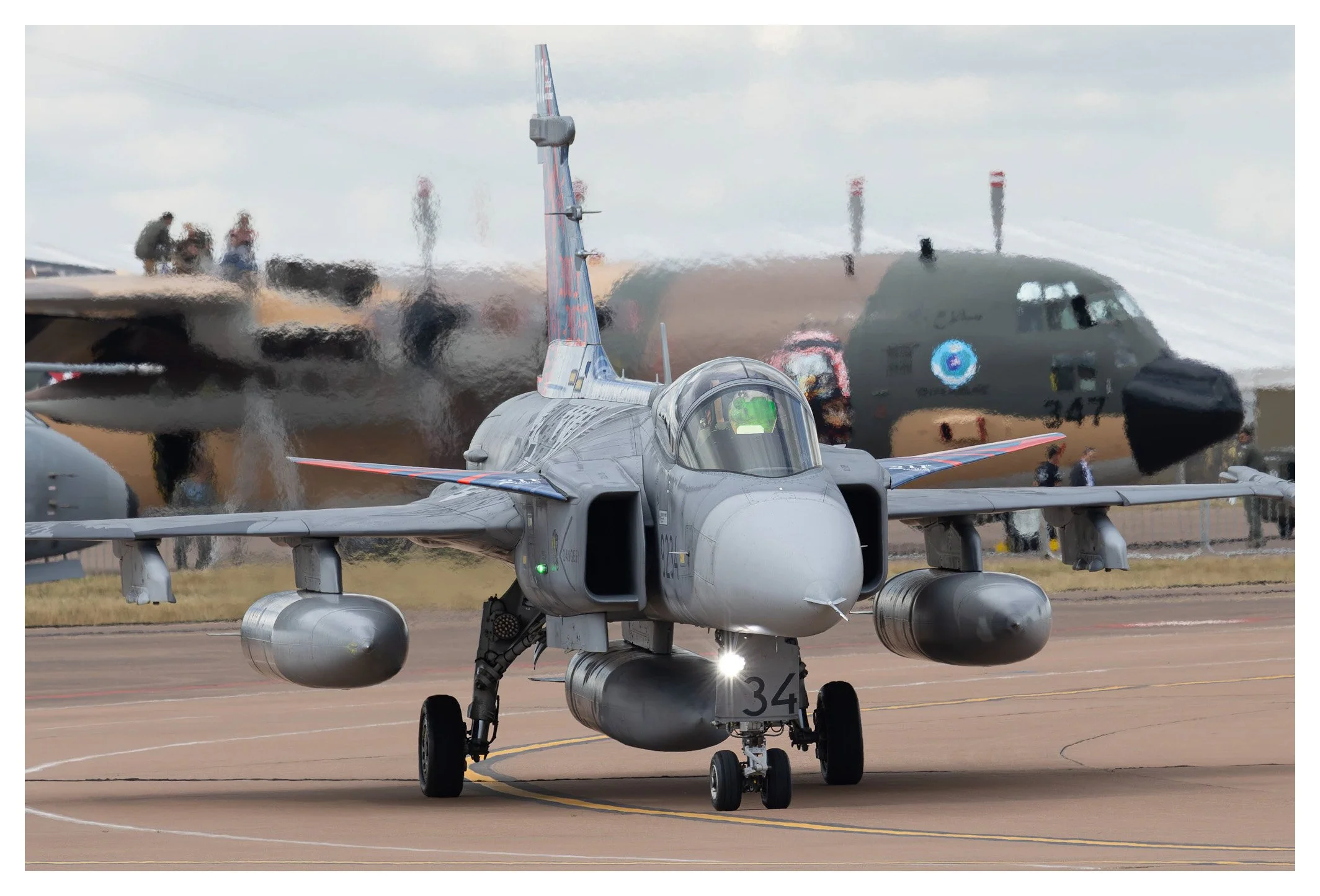 A military fighter jet on the runway at an airshow, with a large transport airplane and several people in the background.