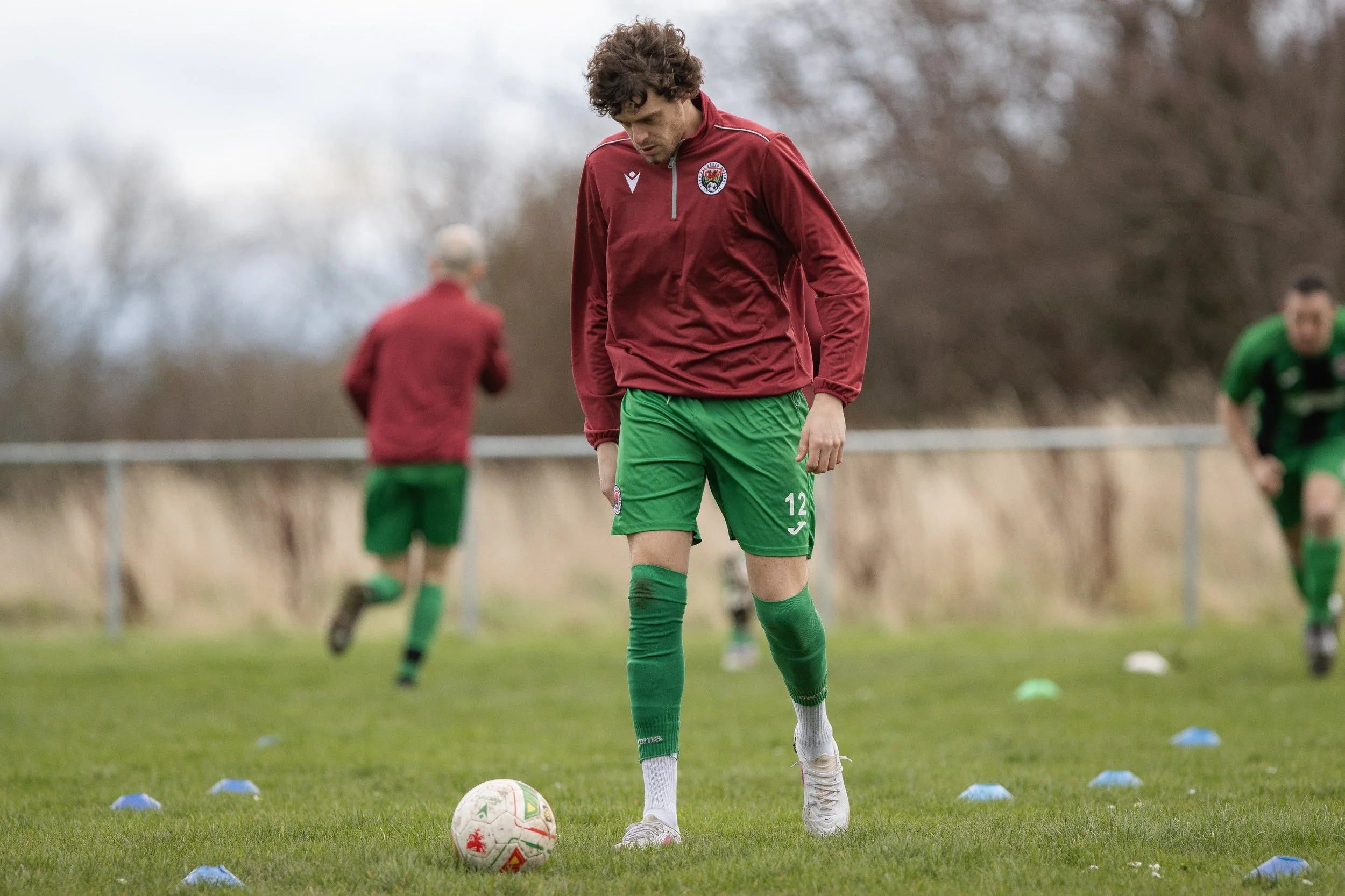 A soccer player in a maroon jacket and green shorts standing on a grass field with a soccer ball at his feet during practice, with two other players in the background, one in a green uniform and the other in maroon, under a cloudy sky.