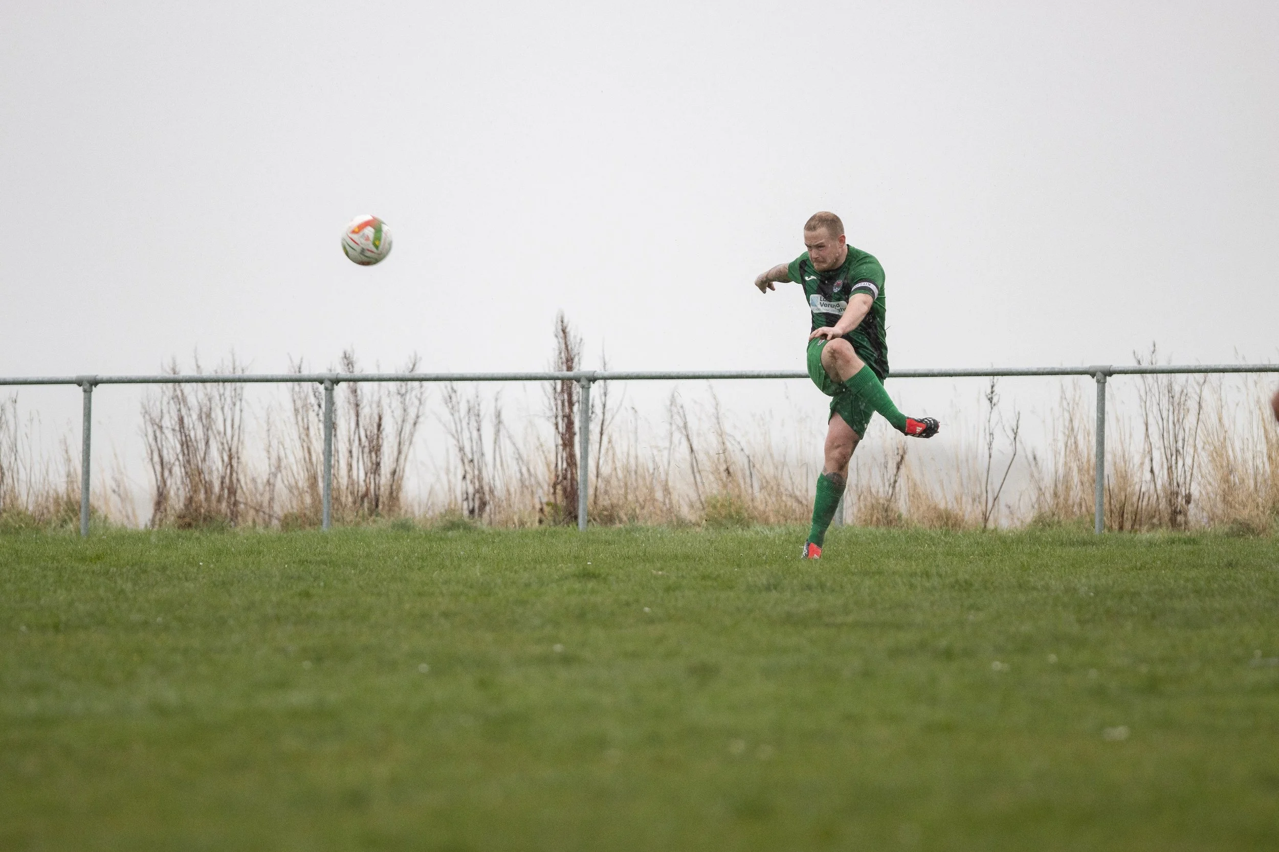 A soccer player in a green and black uniform kicking a soccer ball on a grassy field during a cloudy day.