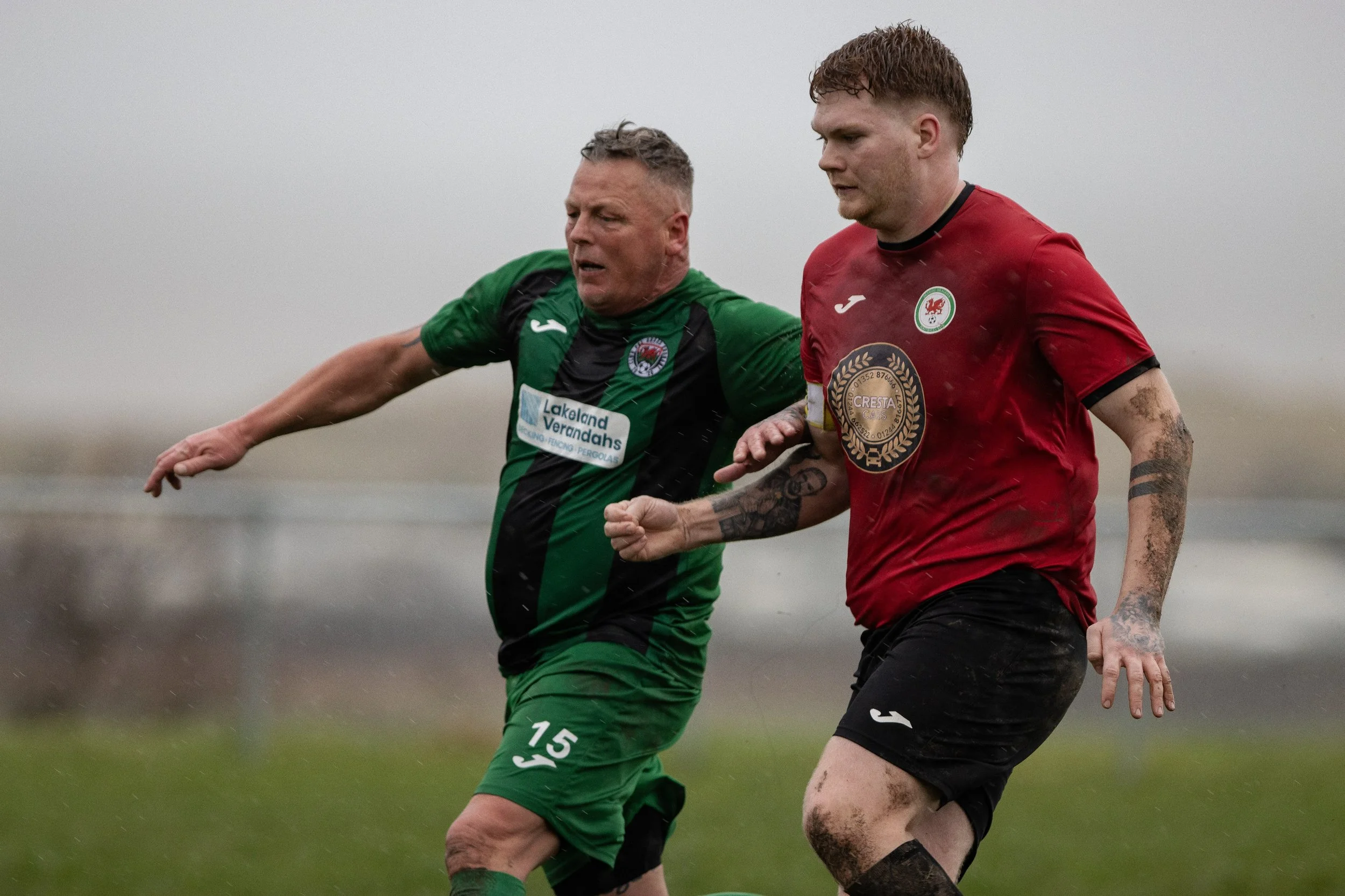 Two soccer players racing for the ball on a muddy field during a rainy day.