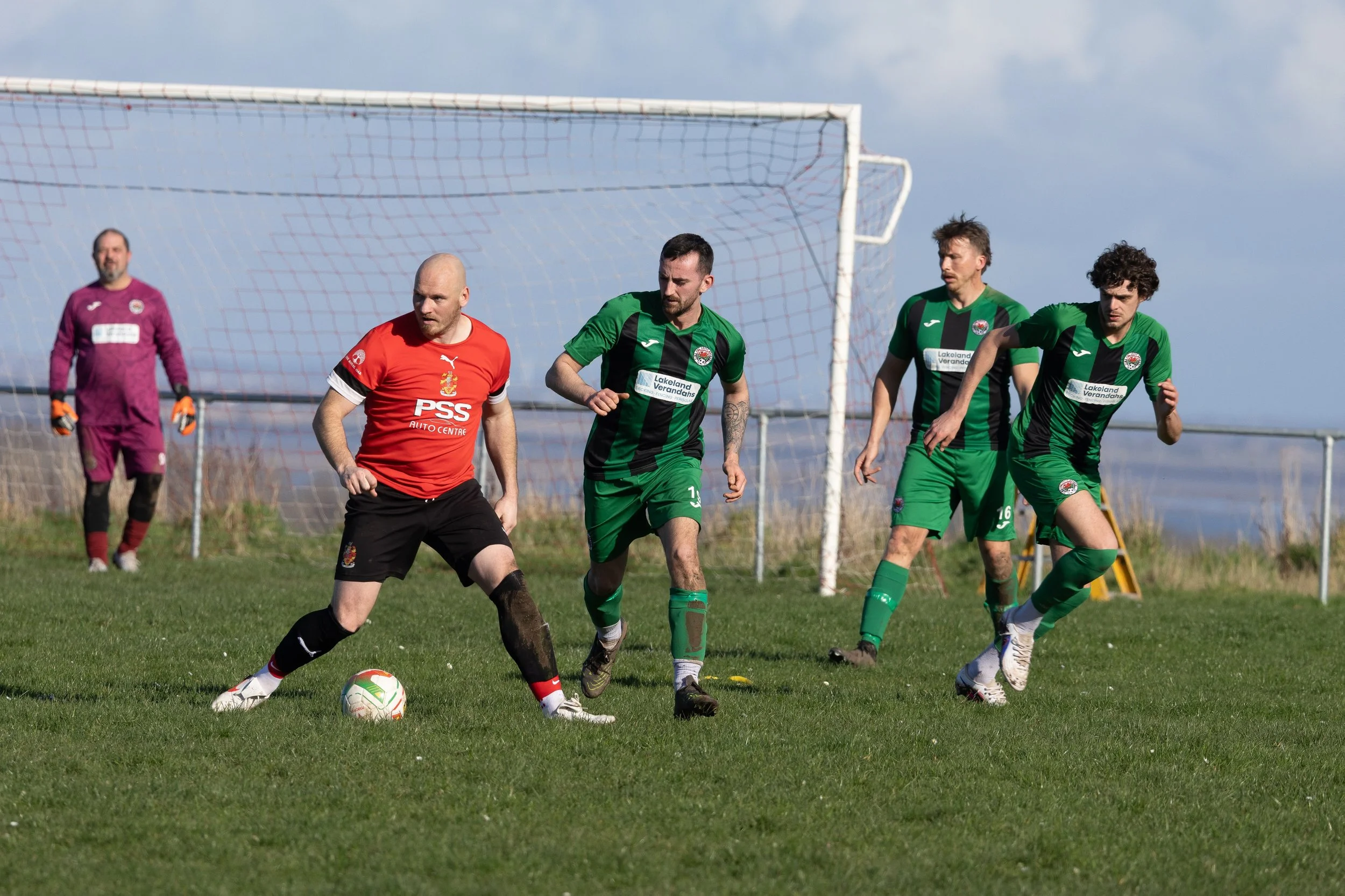 Soccer game with players in green and red uniforms on a grassy field, with a goal and goalie's net in the background under a partly cloudy sky.