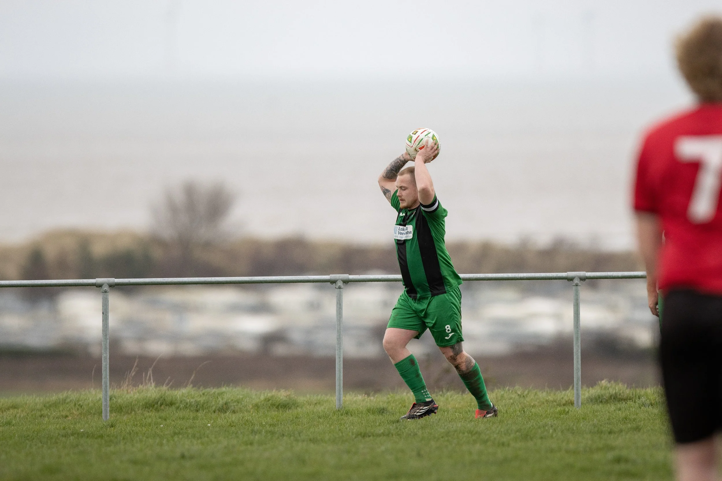 A soccer player in a green uniform prepares to throw the ball during a game, with another player partially visible in a red jersey on the right side.