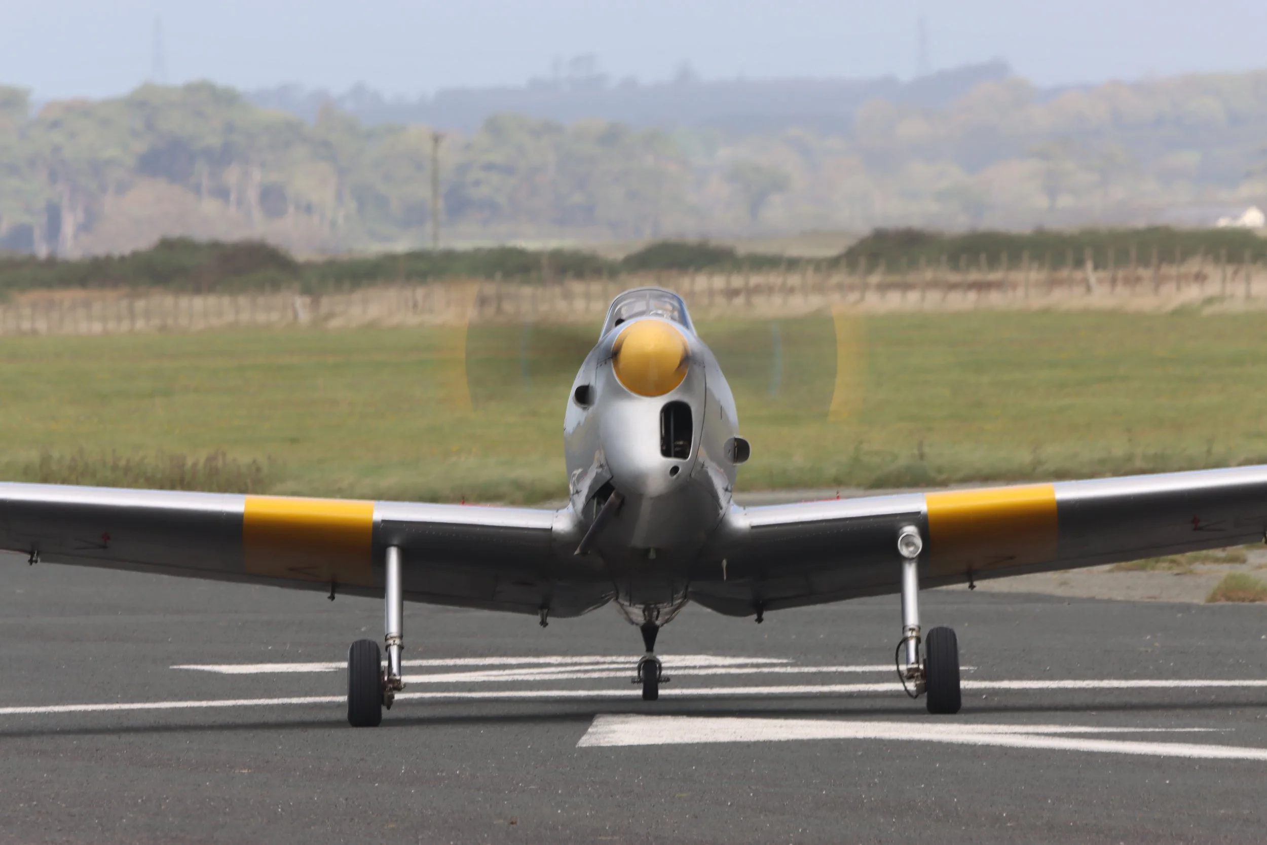 Front view of a silver military jet aircraft with yellow markings, taxiing on runway, with grassy field and trees in background.