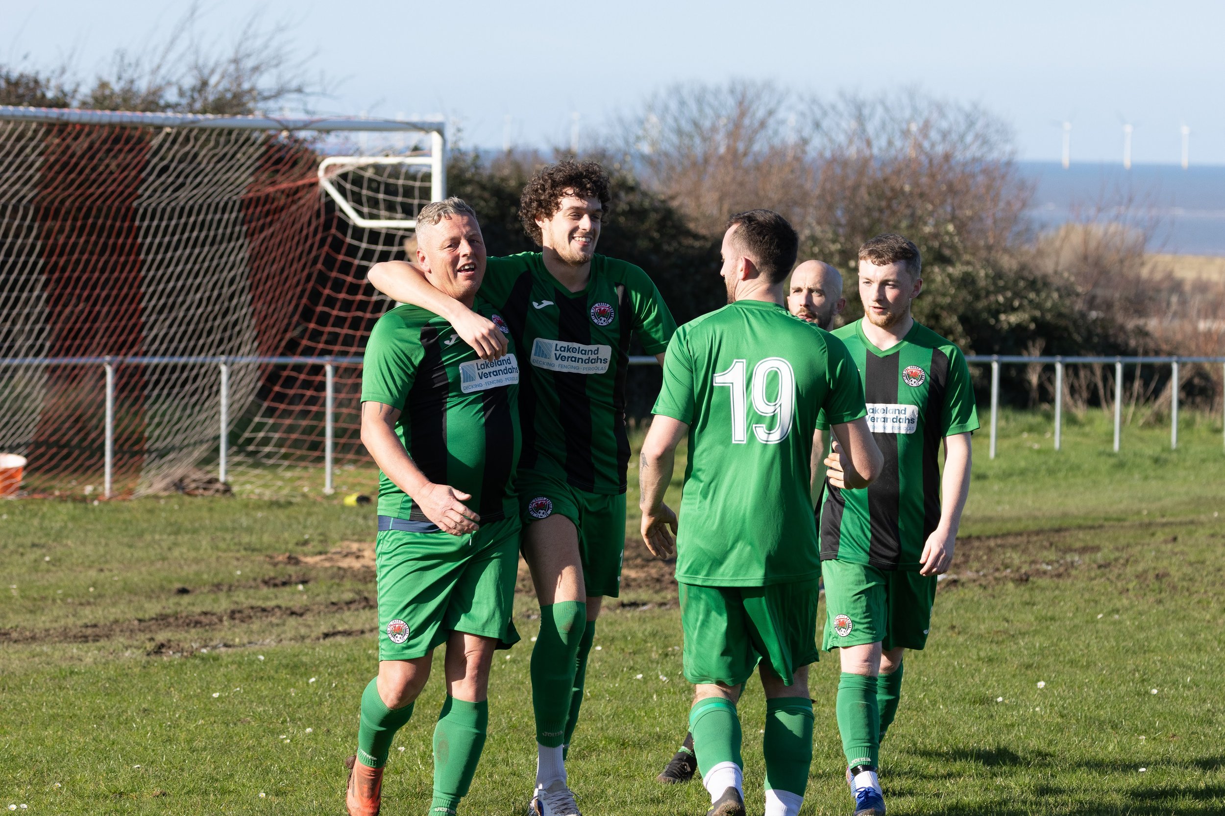 Five soccer players in green and black uniforms celebrating on the field, with one player being embraced by another, during daylight. Soccer goal in background, outdoors with trees and wind turbines visible in the distance.