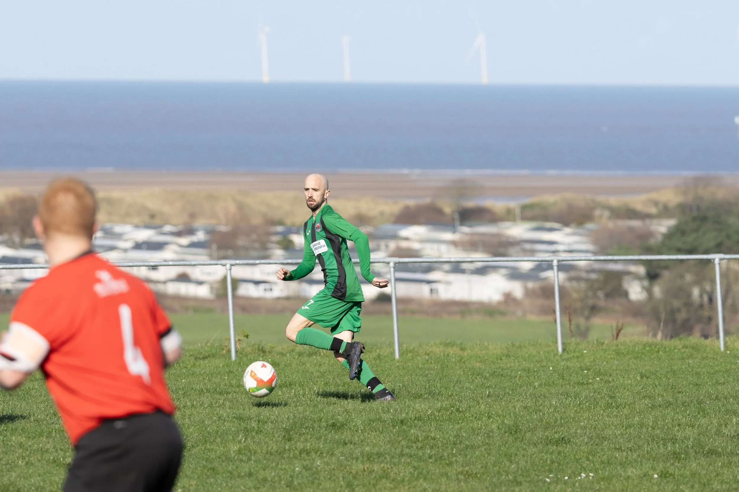 A soccer game in progress on a grassy field, with a player in a green jersey and black shorts about to kick the ball, while another player in a red jersey is in the foreground. In the background, there is a body of water with wind turbines and a dist