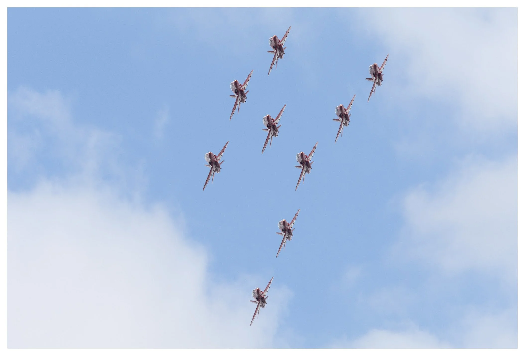 Seven fighter jets performing a synchronized aerobatic formation in a clear blue sky with some scattered clouds.