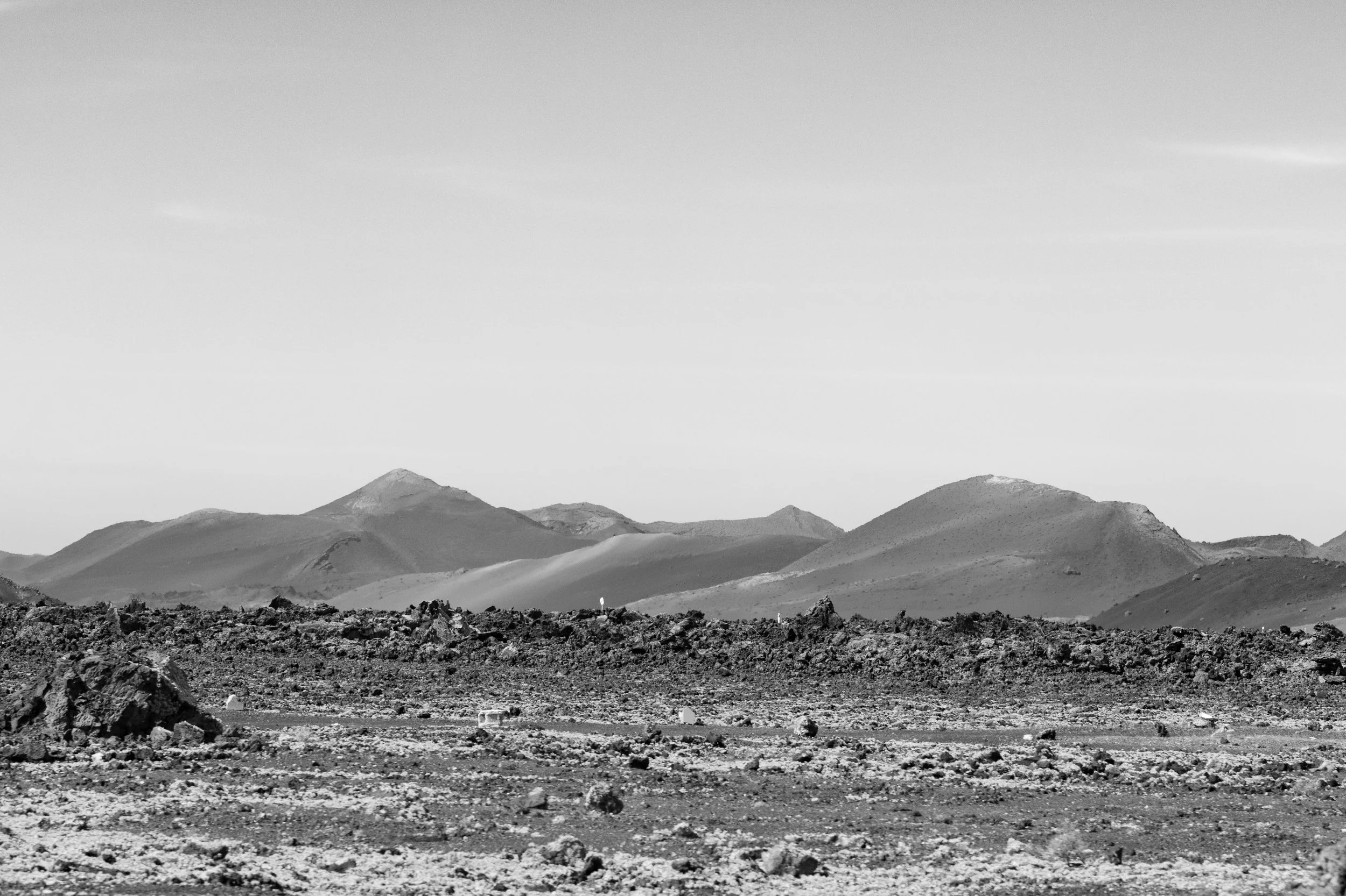 Black-and-white photo of a rocky, barren desert landscape with volcanic hills and mountains in the distance under a clear sky.