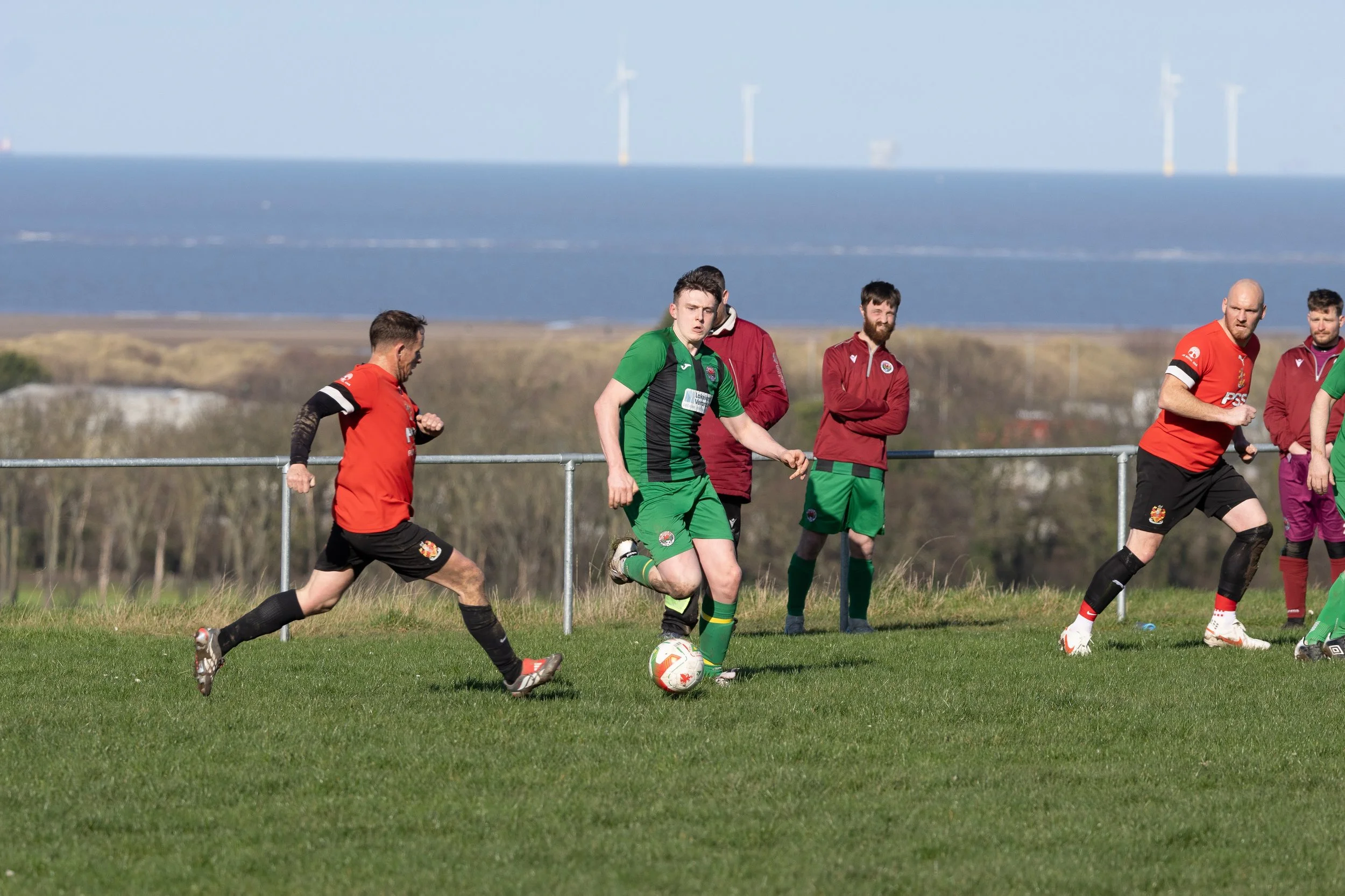 Soccer players in red and green jerseys on a grassy field, with water and wind turbines in the background.