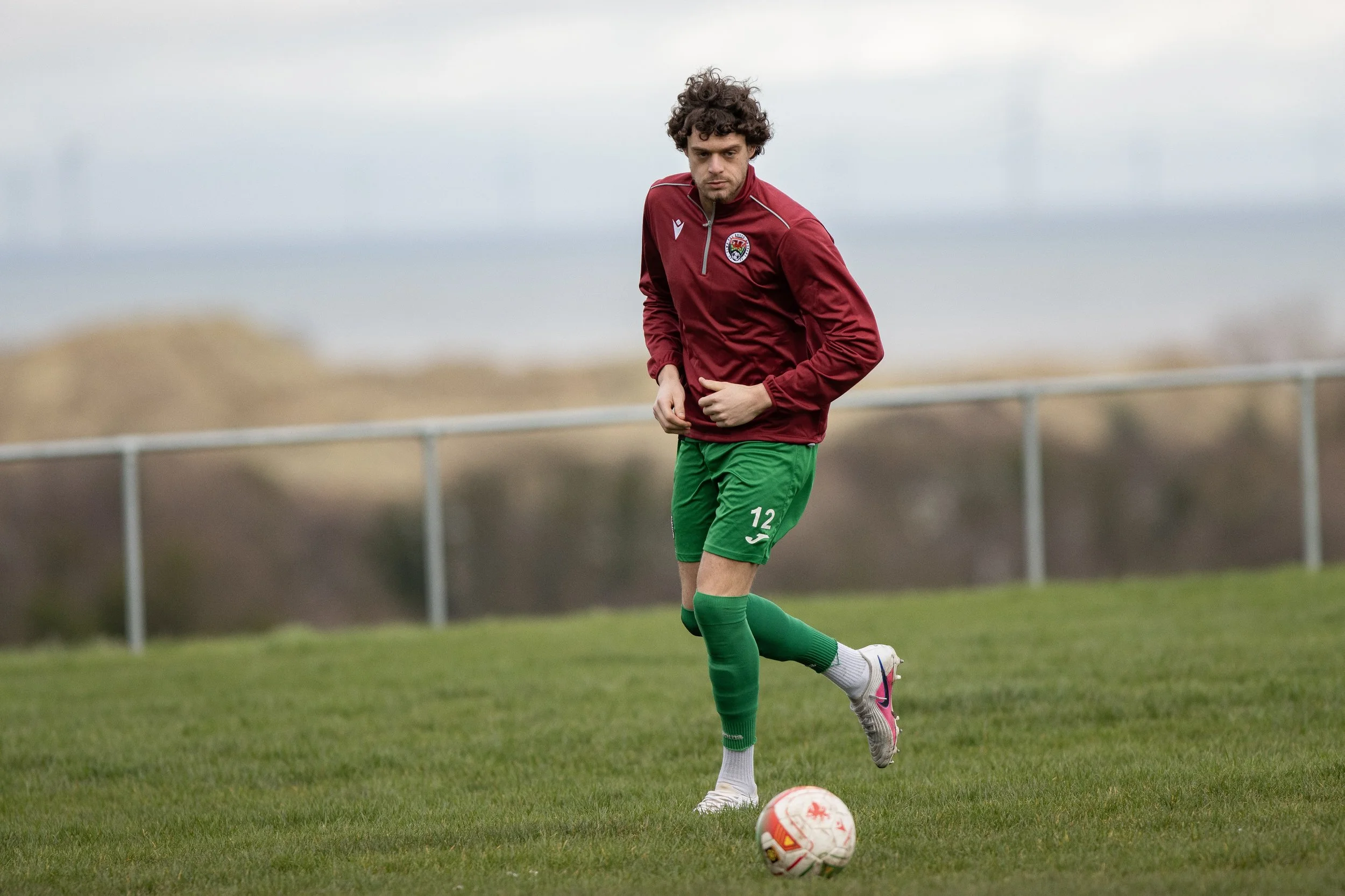 A soccer player wearing a red jacket and green shorts, with the number 12, standing on a grassy field with a soccer ball, under a gray, cloudy sky.