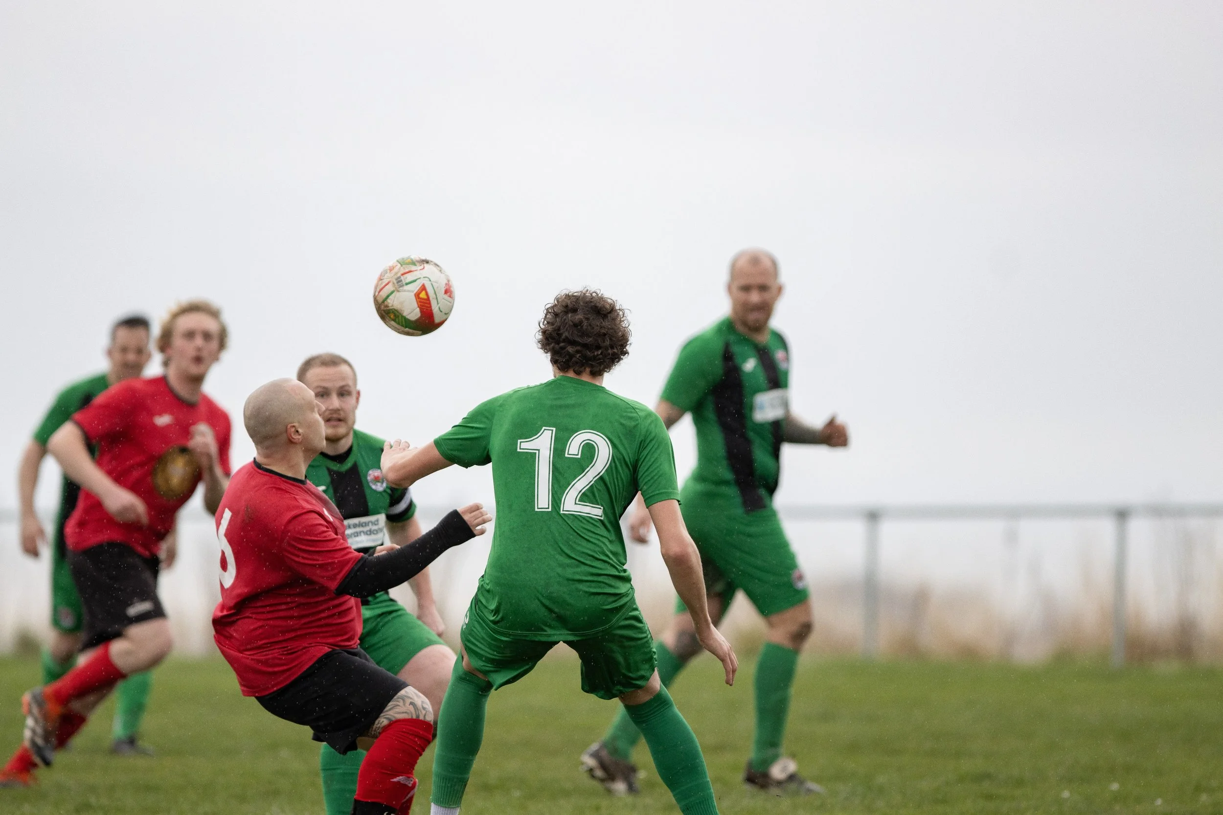 A group of soccer players on the field during a match, with one player in a green jersey marked with the number 12 attempting to head the ball while others, in red and green jerseys, watch or prepare for the next move.