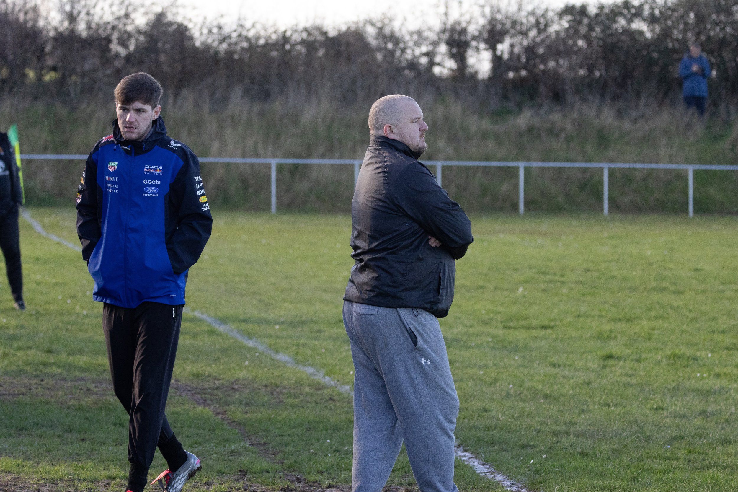 Two men standing on a grassy sports field with a fence and trees in the background, one appears to be a player and the other a coach or spectator.