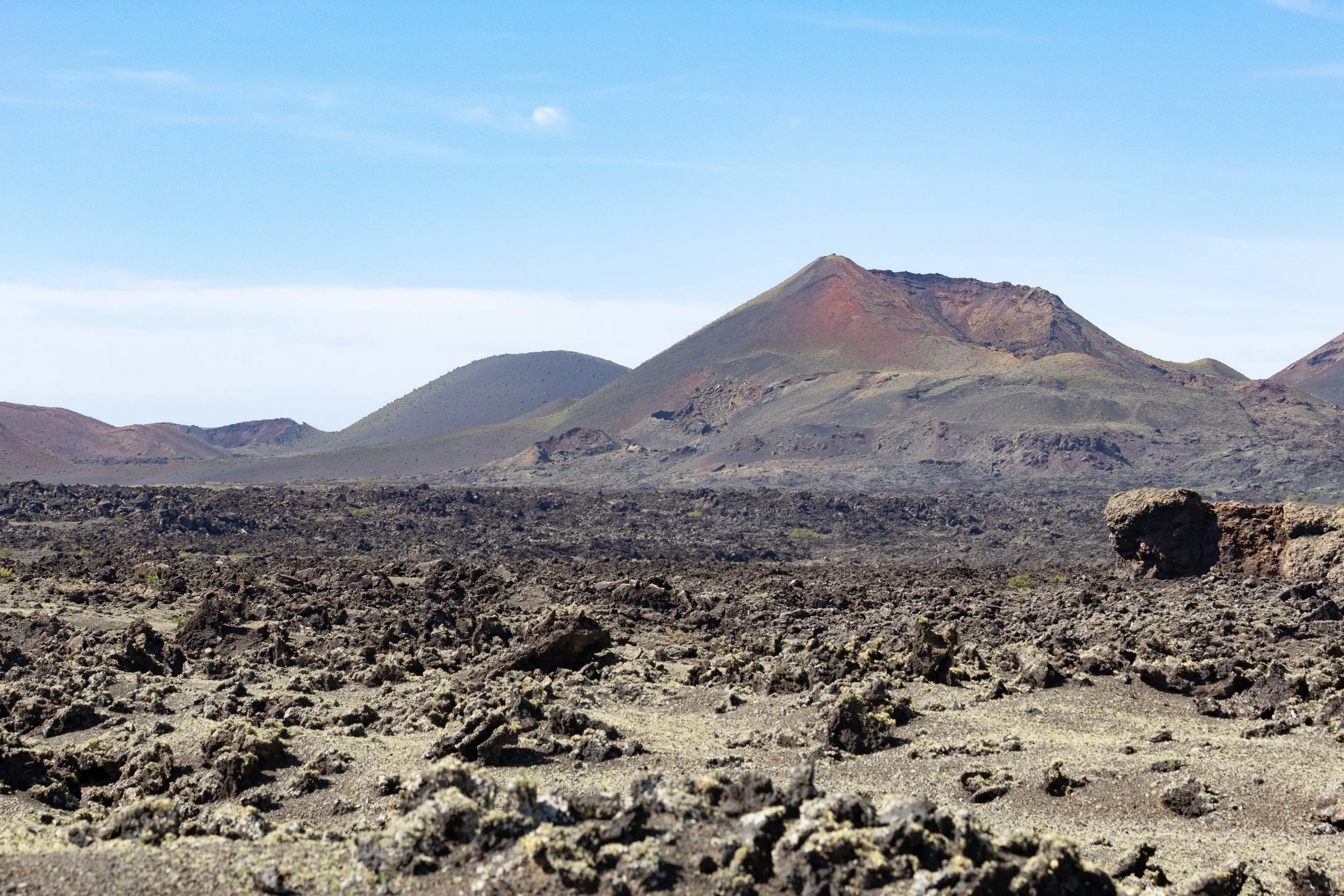A volcanic landscape with dark, rugged lava rocks in the foreground and distant volcanic mountains under a blue sky.