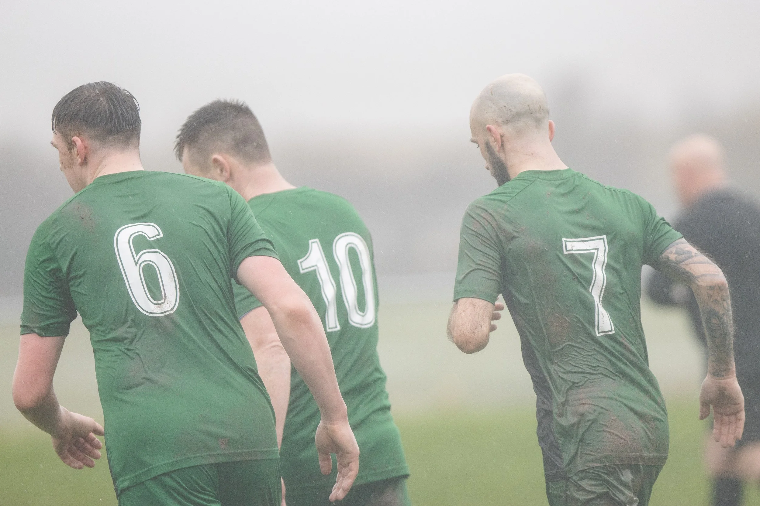 Four soccer players in green jerseys walking on a foggy field, their backs turned to the camera, with numbers 6, 10, and 7 visible.
