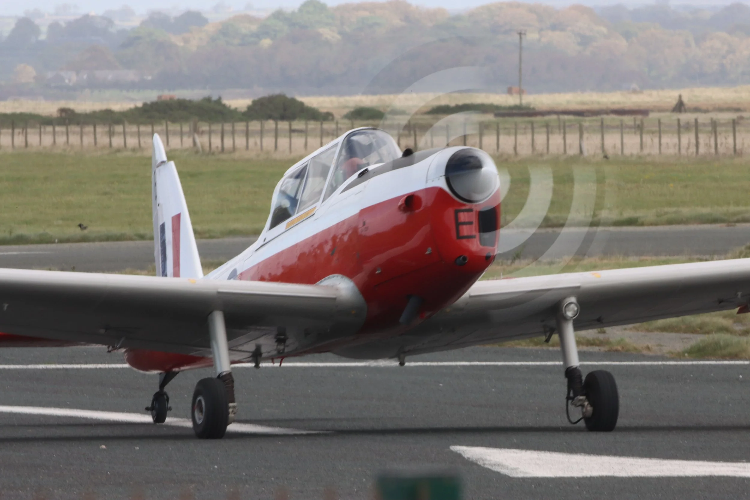 A small vintage aircraft with a red and white exterior on a runway during daytime, with a rural landscape in the background.