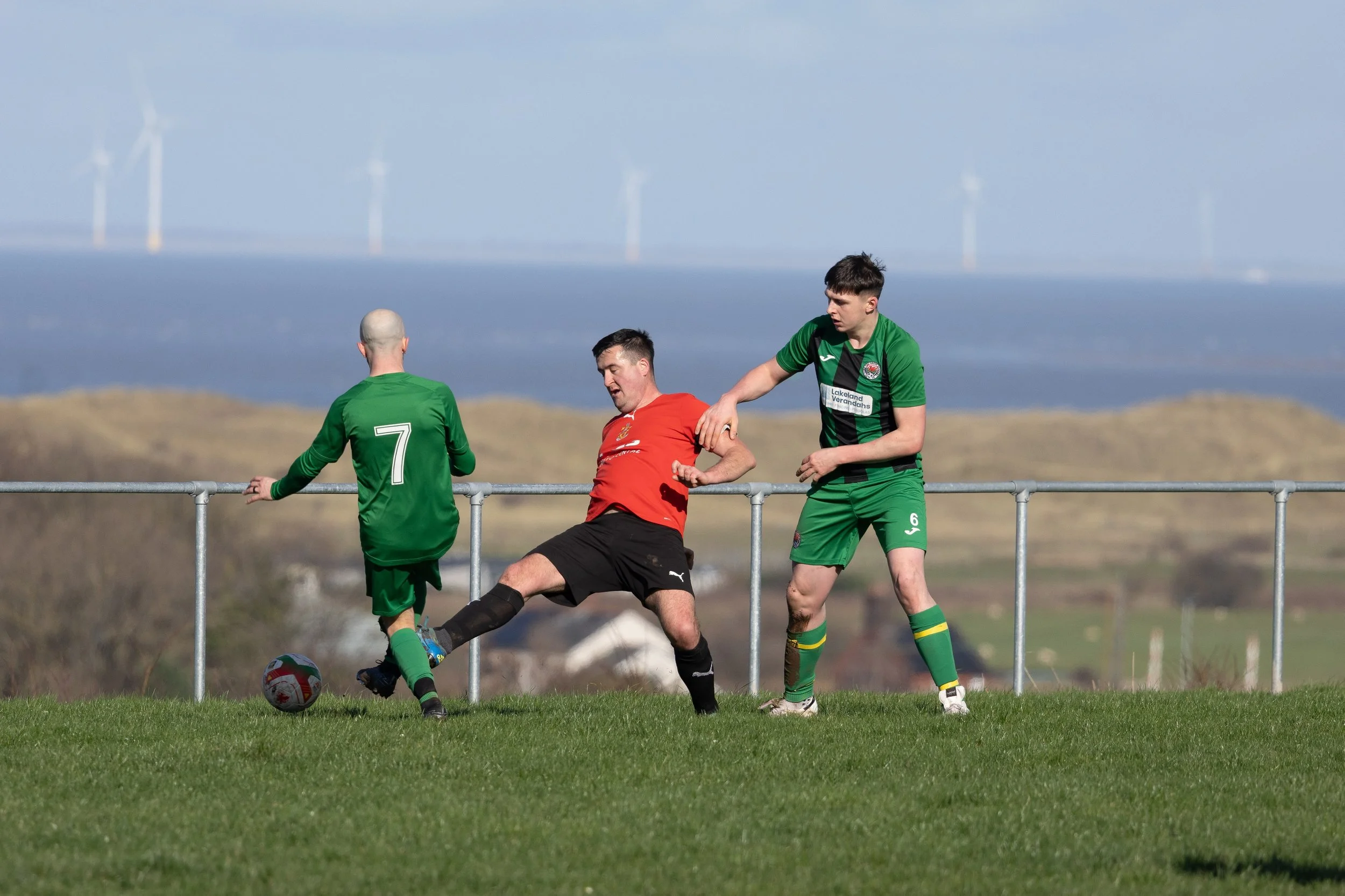 Three soccer players on a field during a game, two in green uniforms and one in a red shirt, with a scenic landscape and wind turbines in the background.