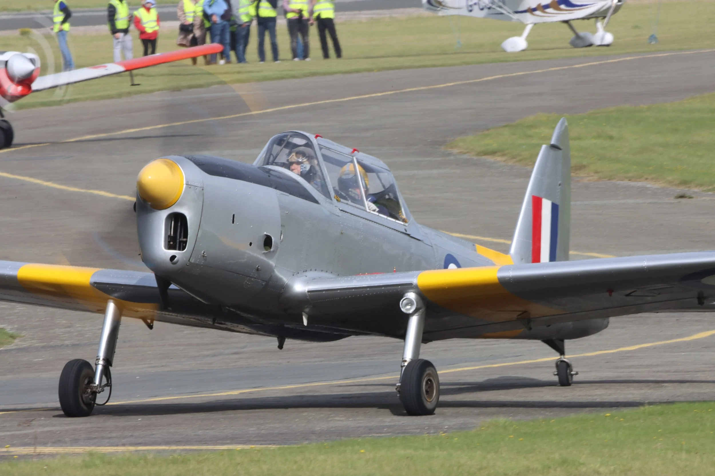 A vintage military jet aircraft on the ground with a pilot inside, and a group of people watching in the background.