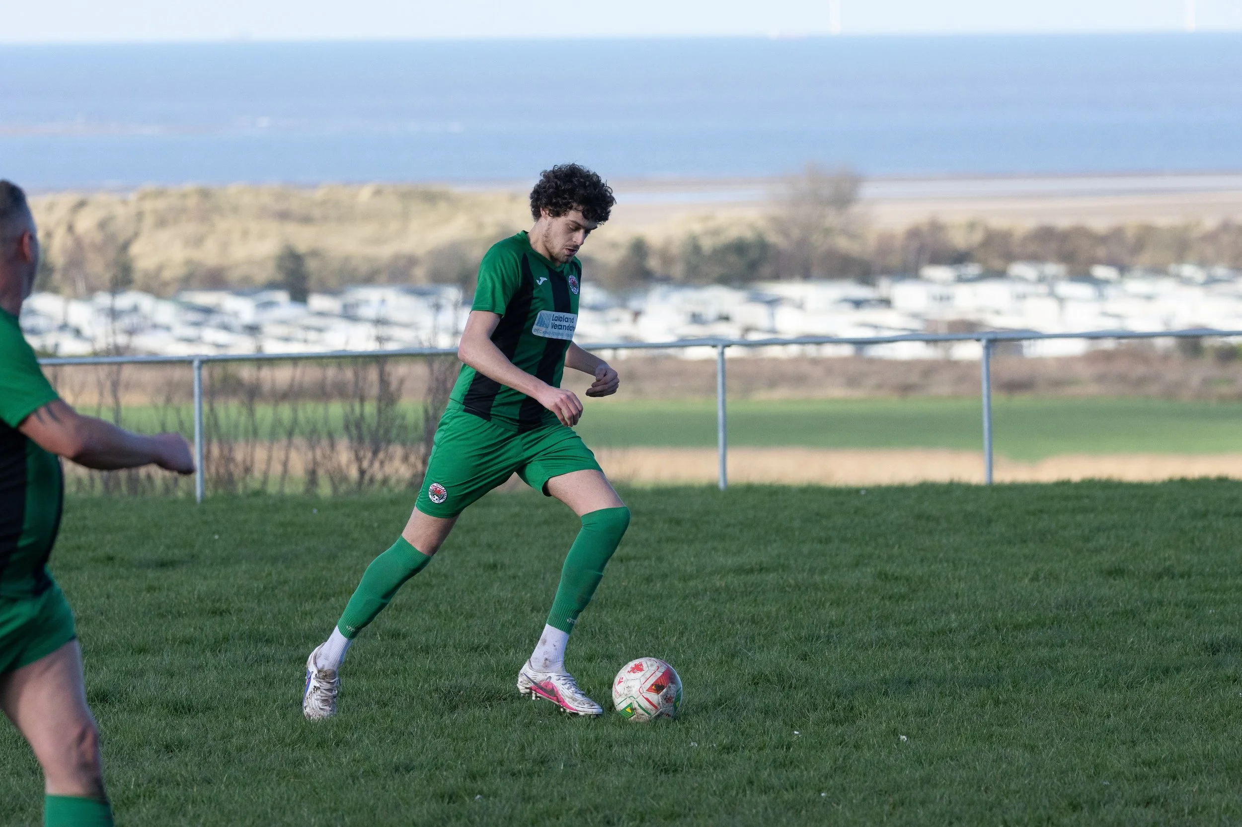 A soccer player in a green and black uniform is about to kick a soccer ball on a grassy field, with a scenic background of fields, trees, and water.
