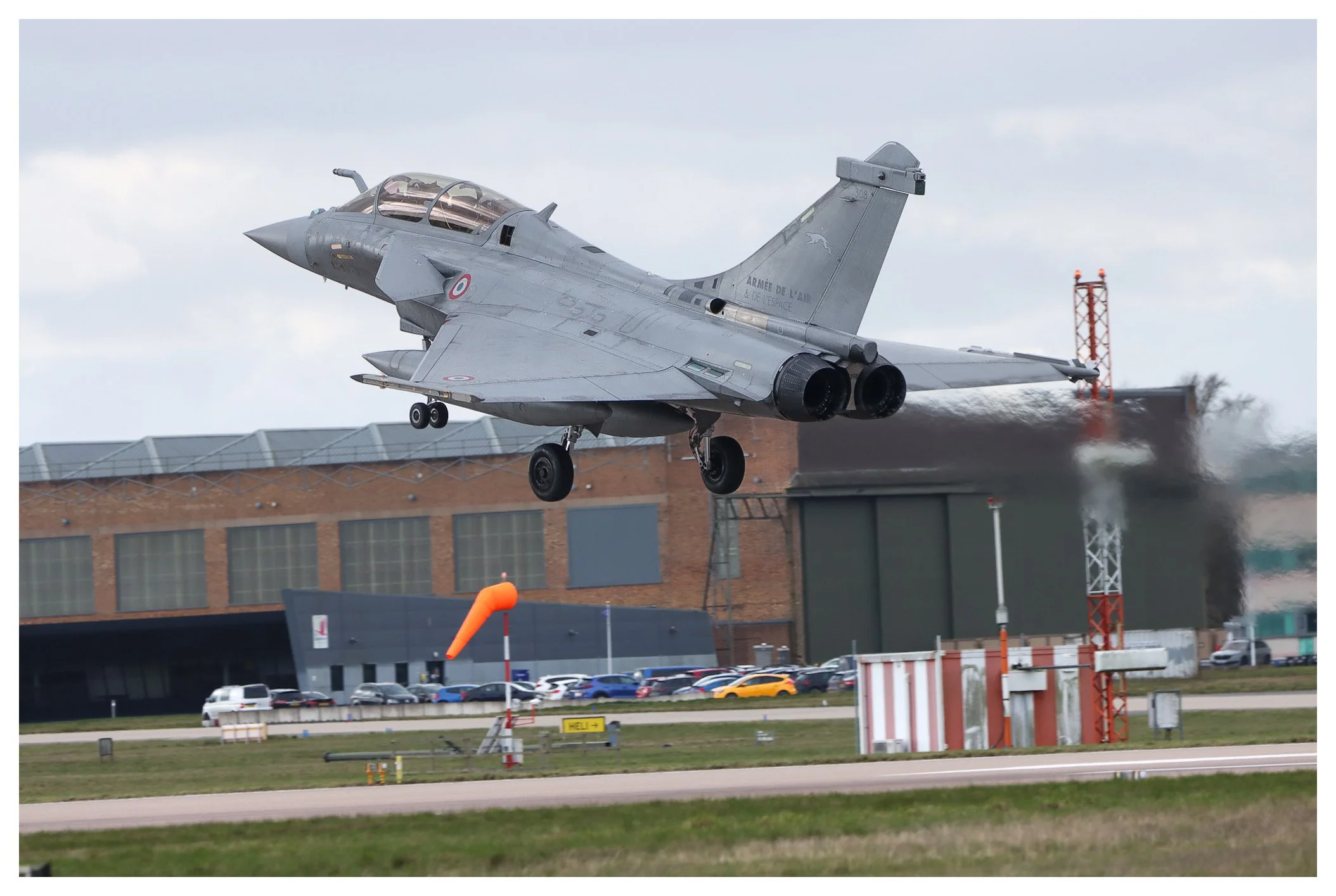 A French military fighter jet, specifically a Dassault Rafale, is taking off from a runway during daytime. The aircraft is in mid-air just above the ground, with its landing gear still extended. The background features an airport building, parked car