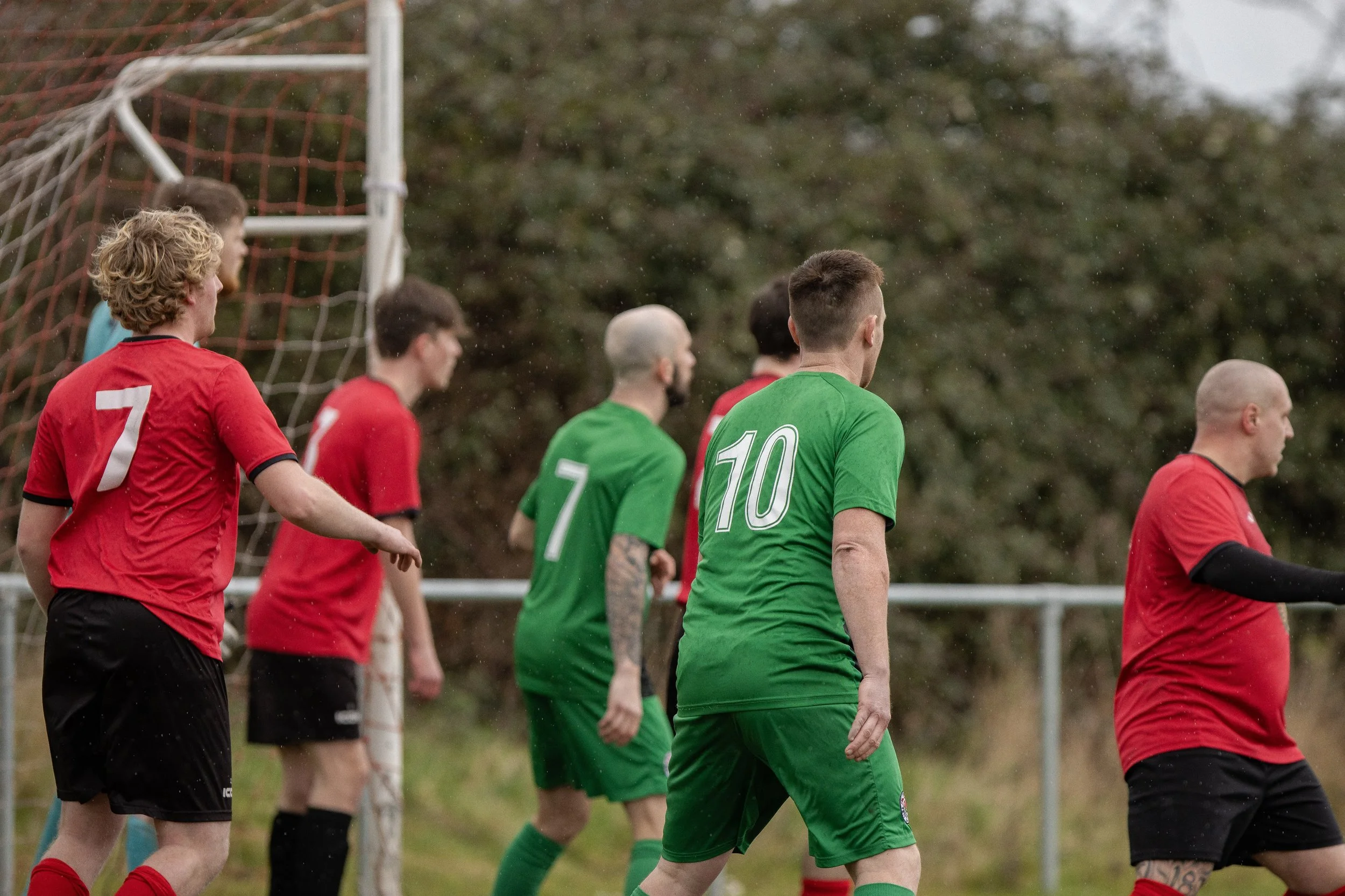 Soccer players in red and green uniforms on the field near a goal post, likely during a match on a cloudy day with trees in the background.