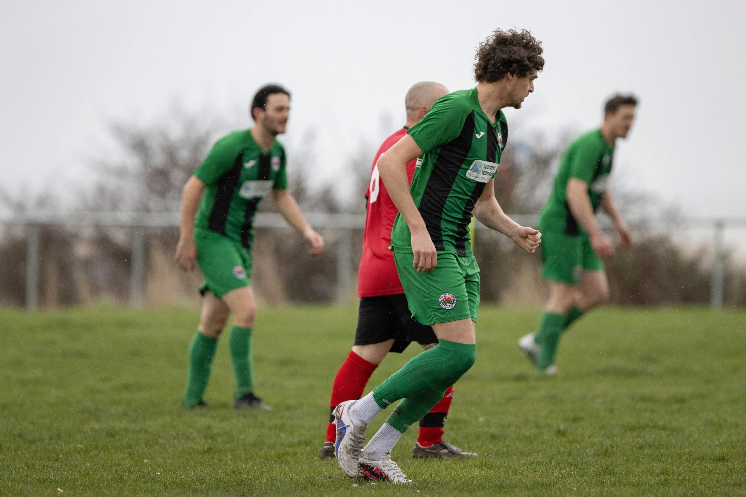 Soccer players on a grassy field, wearing green and black uniforms, participating in a match on a cloudy day.