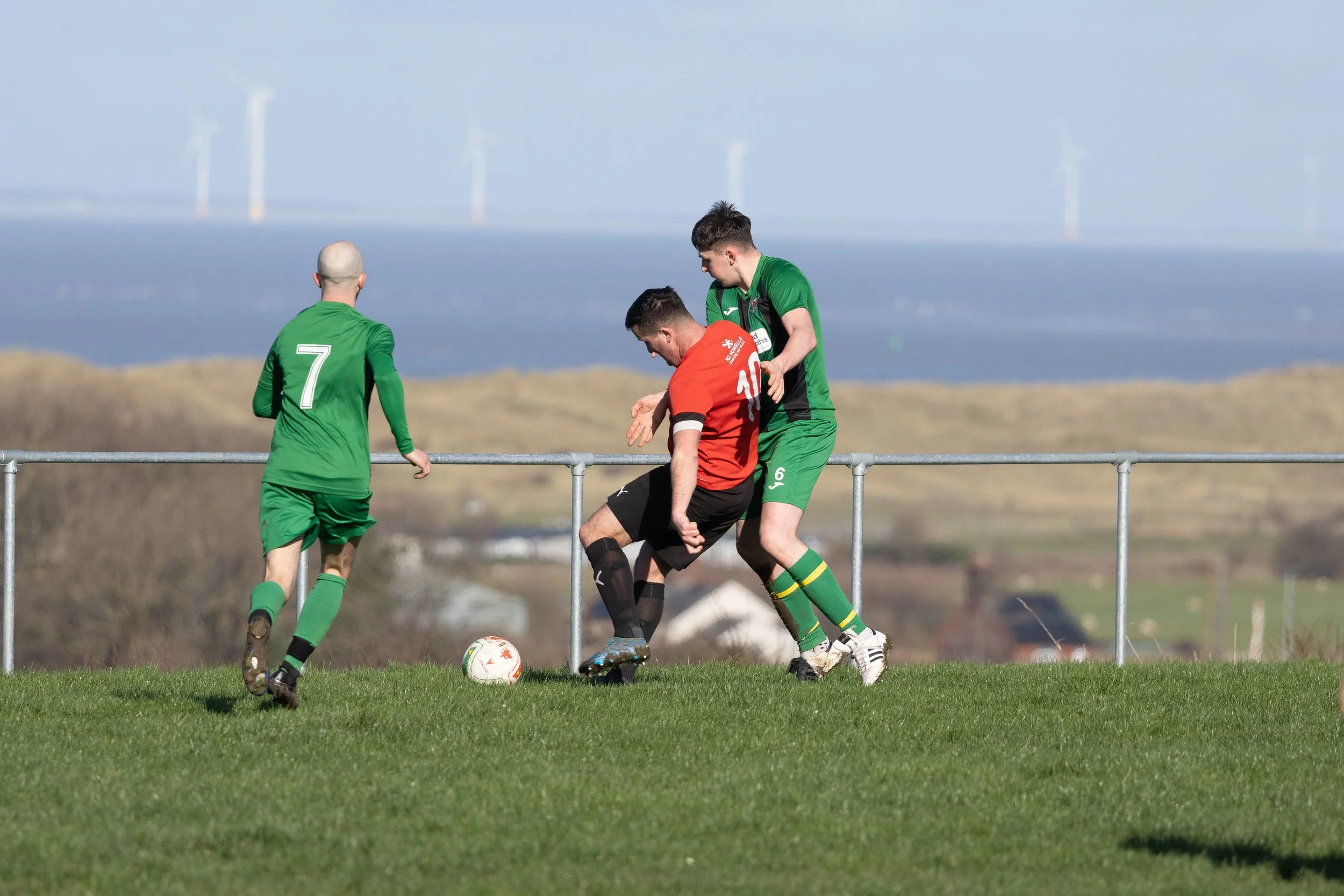 Soccer players in green and red jerseys competing for the ball on a grassy field, with a rural landscape and wind turbines in the background.