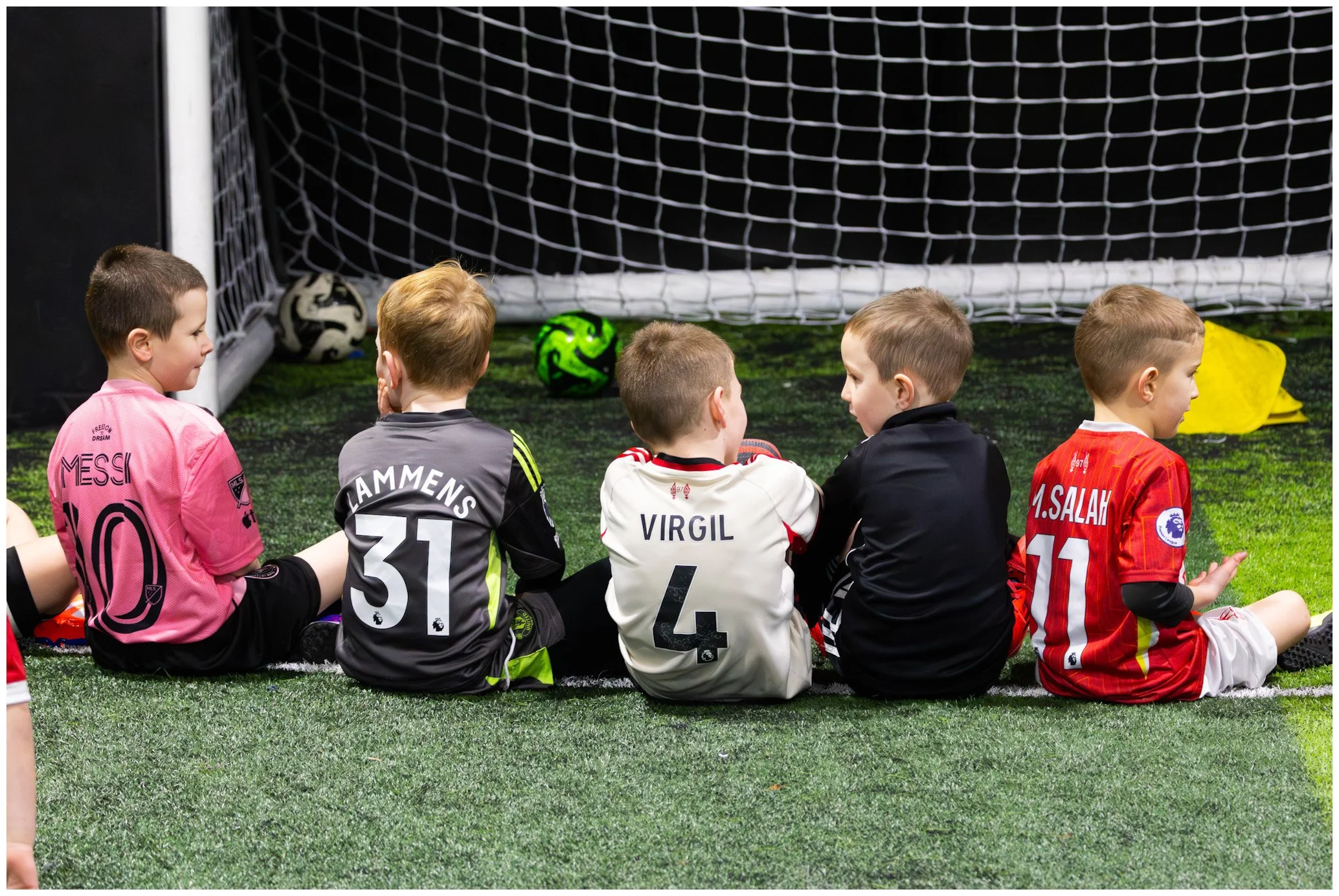 Five young boys sitting on the field near a soccer goal, wearing team jerseys, with soccer balls behind them.